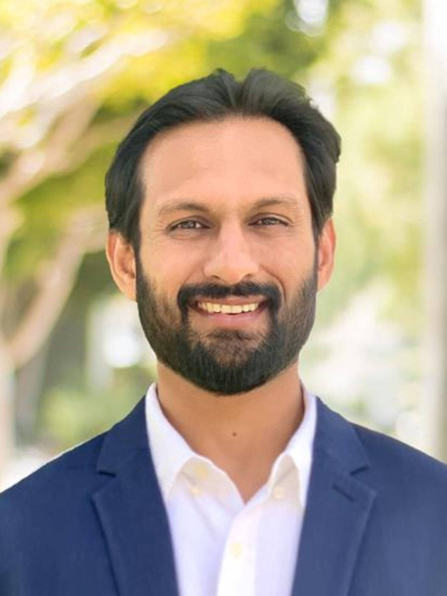 A man with a beard and dark hair, wearing a white shirt and blue blazer, smiles at the camera. The background is blurred, showcasing green foliage and soft sunlight, evoking curiosity about Genea's serene setting.