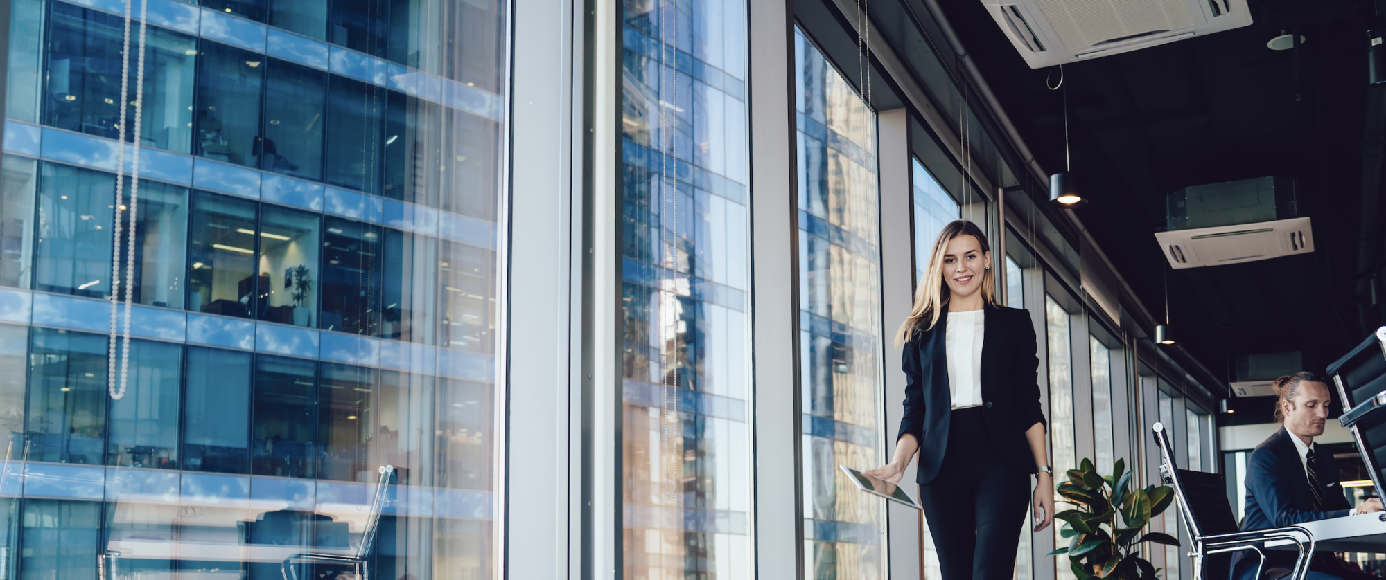 A woman in a black suit walks confidently through a modern office with floor-to-ceiling windows, embodying the poise of seasoned security consultants. She holds a tablet and smiles while, in the background, a man works at a desk. The cityscape of tall buildings stretches beyond.