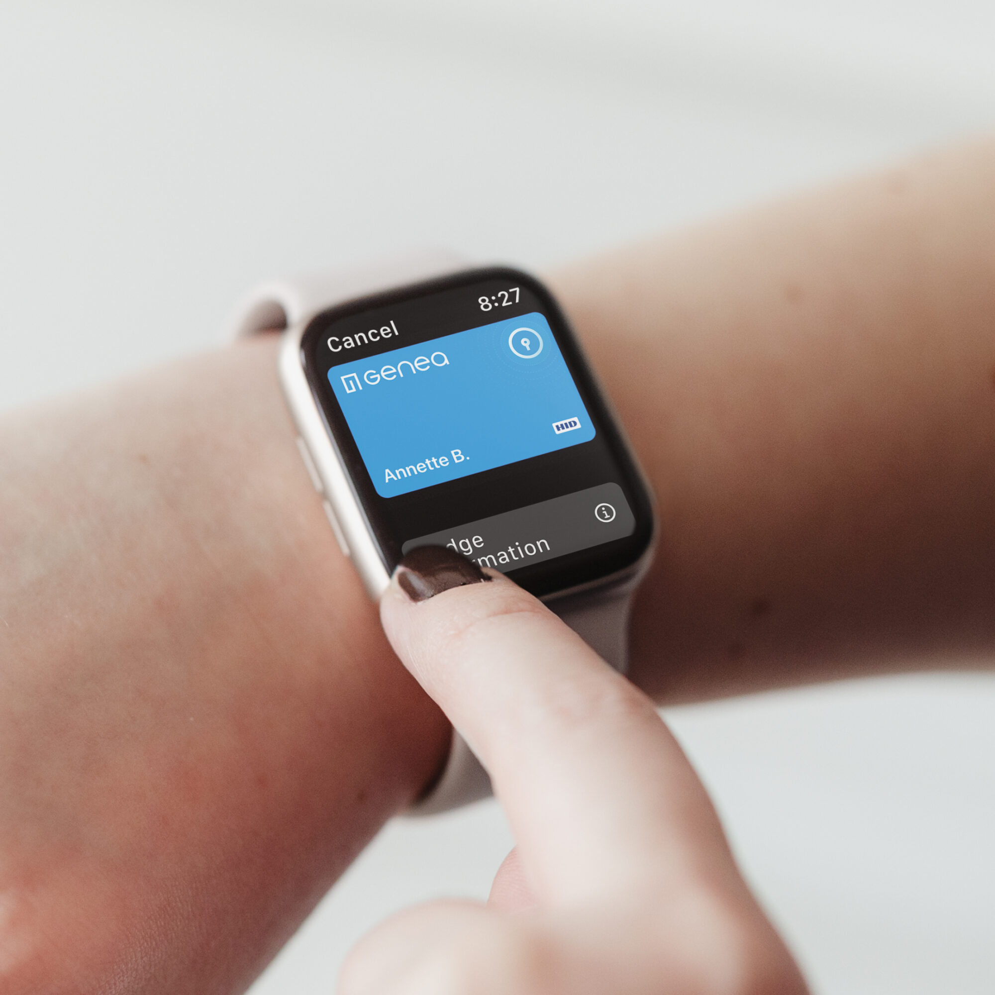 Close-up of a smartwatch on a person's wrist displaying a blue screen with the title Genea and name Annette B. The person is interacting with the watch, possibly accessing Apple Wallet, and the time shown is 8:27.