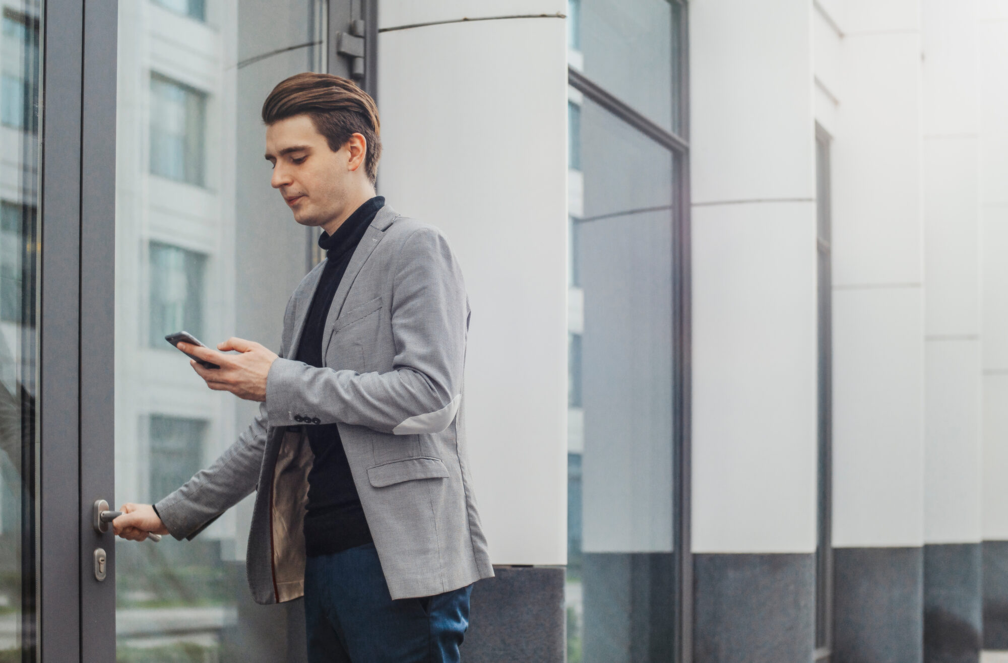 A person wearing a gray blazer and dark turtleneck holds a smartphone while opening a glass door with the other, equipped with magnetic locks. They stand outside a modern building featuring reflective windows.