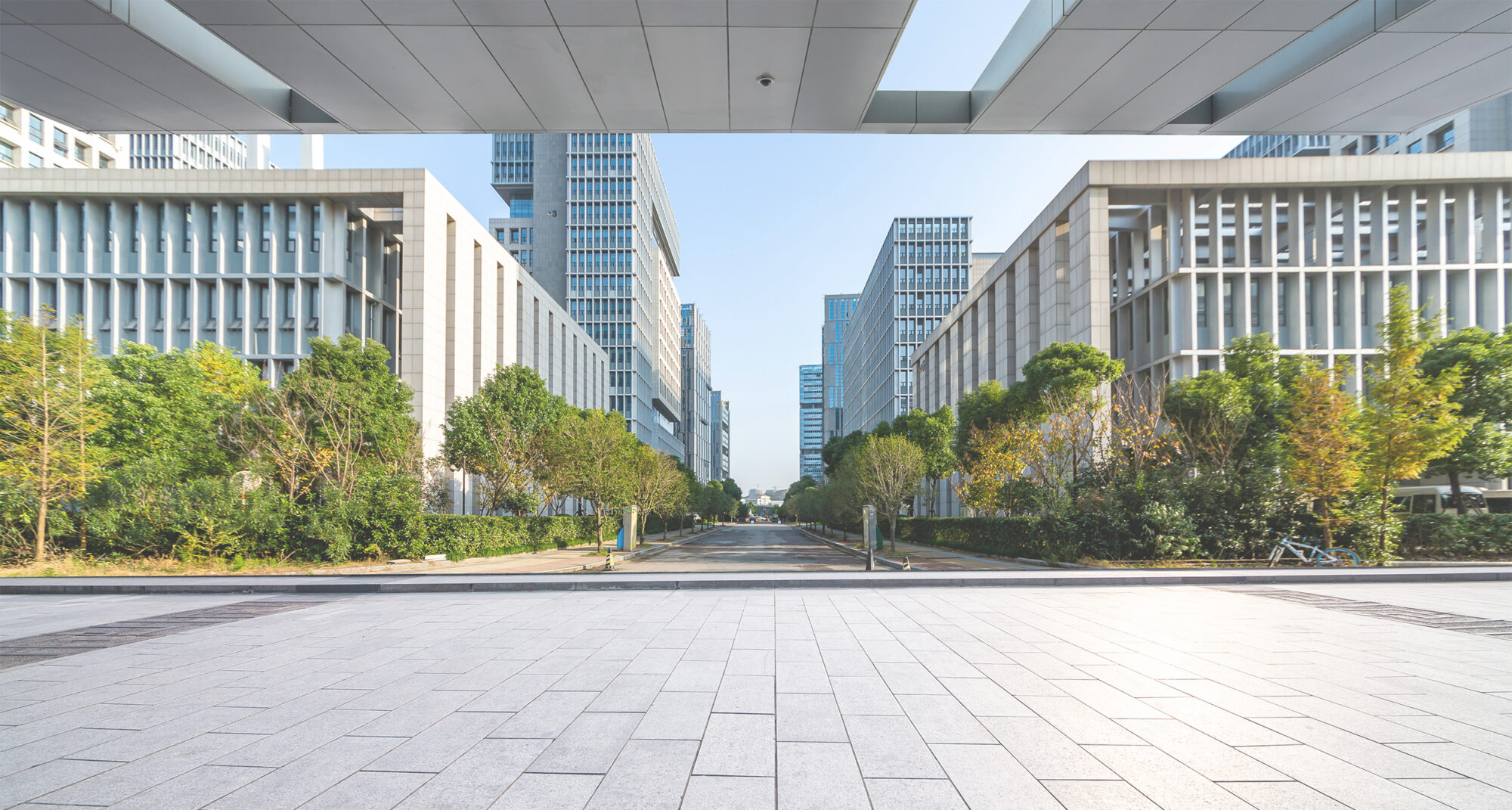 A wide-angle view of a modern urban landscape, showcasing a pathway flanked by trees and contemporary buildings with gridded facades synonymous with innovative IT hubs. The scene is framed by an overhead structure, underlined by a clear blue sky in the background.