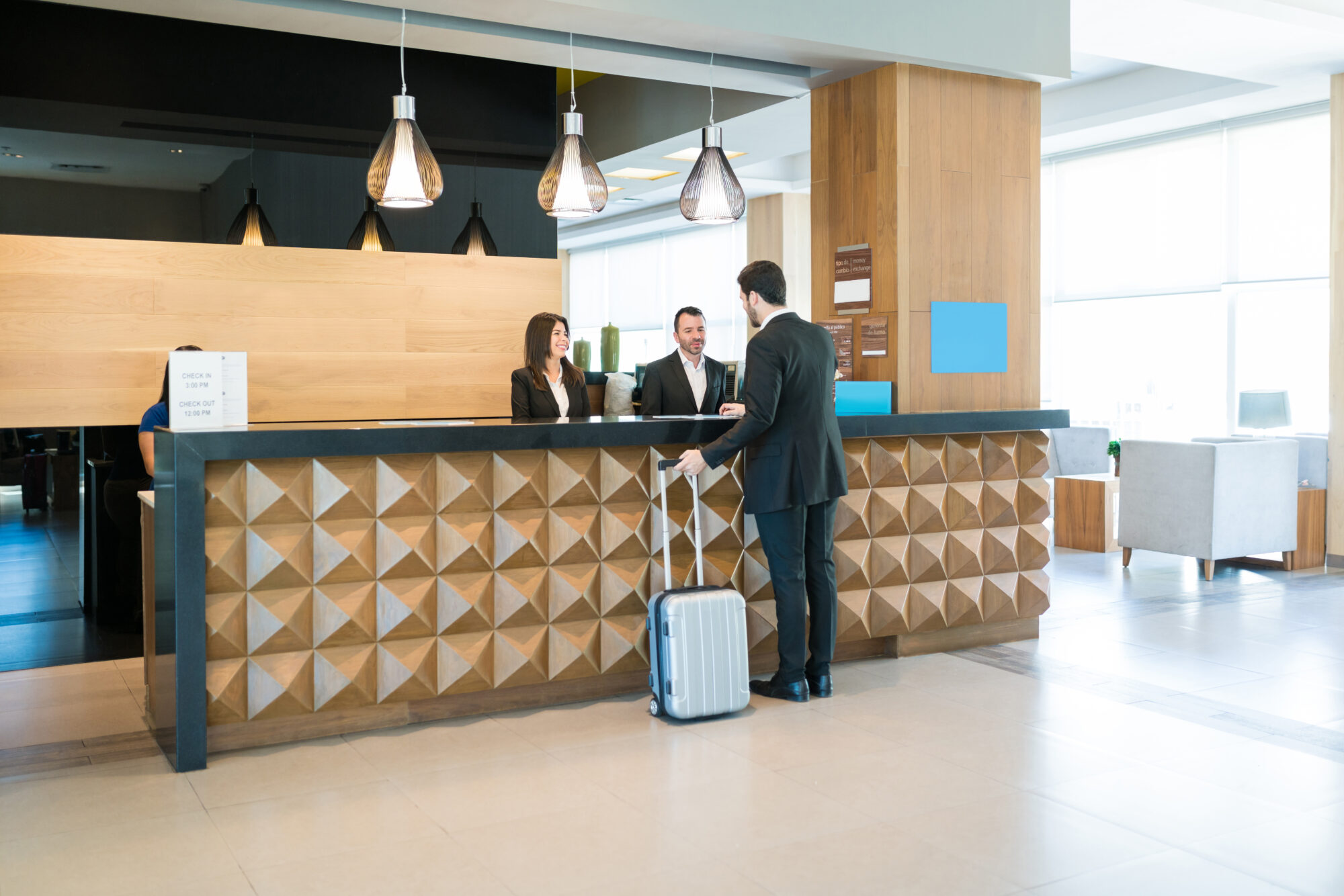 A man in a suit with a suitcase checks in at a modern hotel reception desk equipped with an advanced access control system. Two staff members, a man and a woman, assist him as the geometric paneling and contemporary lighting enhance the welcoming atmosphere.