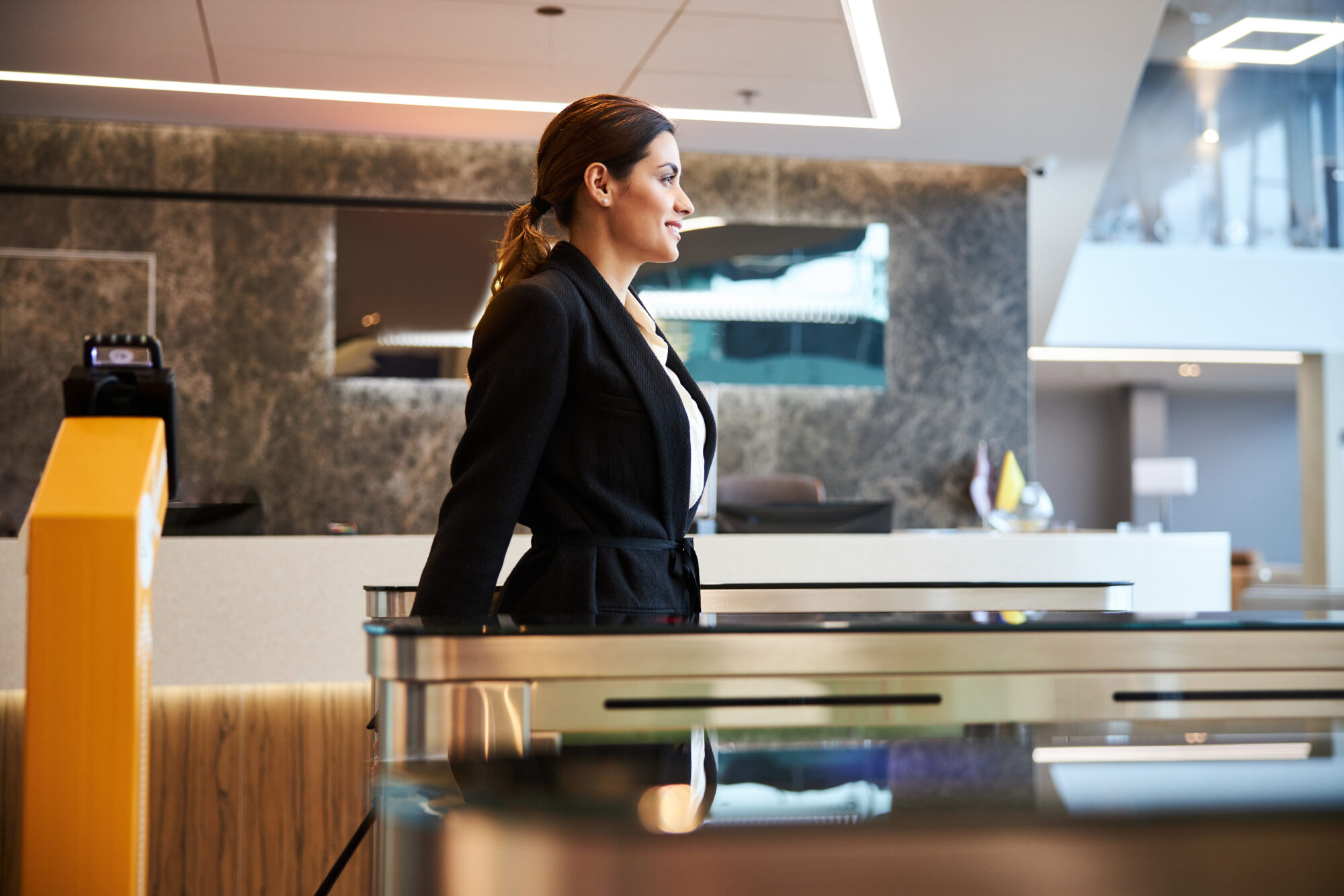 A woman in a black blazer stands at a modern reception desk in a sleek, contemporary office lobby. She appears to be waiting or discussing information security with someone out of frame. The area is well-lit with stylish decor and a reflective surface behind her.