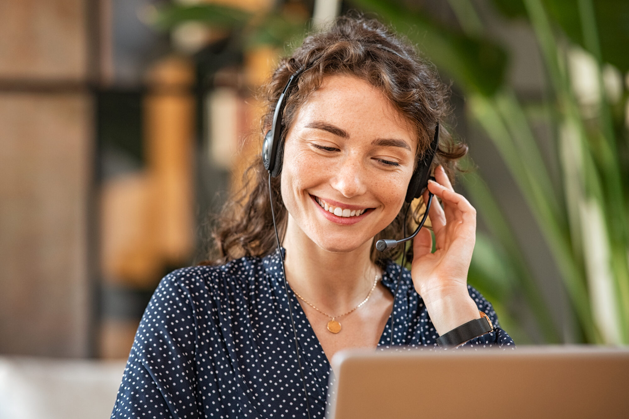 A woman with curly hair is smiling while wearing a headset and looking at a laptop, possibly managing her finances through Apple Wallet. Dressed in a dark polka dot shirt, she sits in a bright room with green plants in the background.