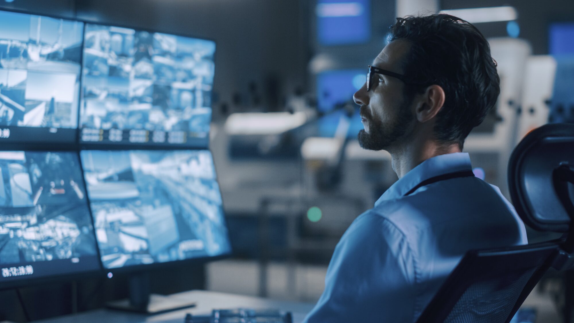 A person wearing glasses is seated in a dimly lit room, monitoring multiple Ava Security camera feeds displayed on large screens. The scene conveys a sense of focus and attentiveness, suggesting a sophisticated security or surveillance control center.