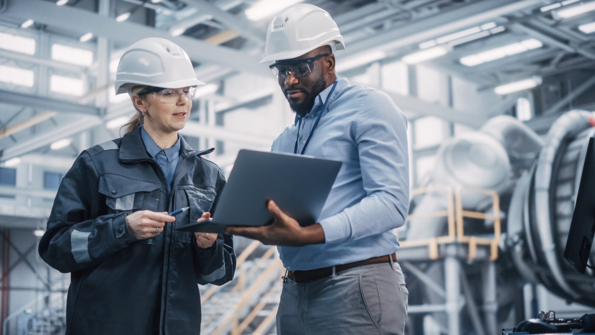 Two building engineers wearing hard hats and safety glasses engage in discussion. One holds a laptop, while the other gestures with a pen. They're in an industrial setting, surrounded by machinery and a mezzanine in the background, focused on their latest project assessment.