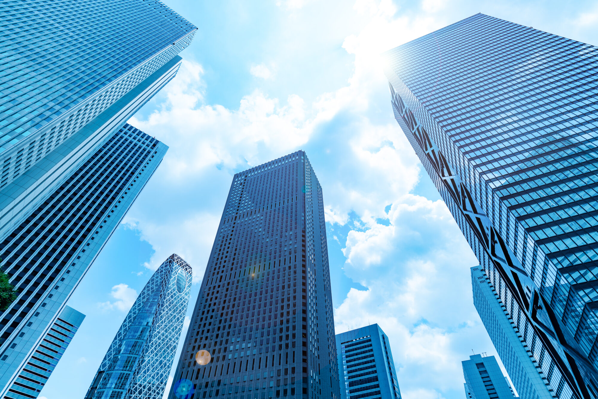 A view looking up at several modern skyscrapers against a bright blue sky with fluffy clouds. Sunlight shines from the upper right corner, reflecting off the glass and steel surfaces of these IT hubs, embodying the cutting-edge security and innovation thriving within their towering heights.