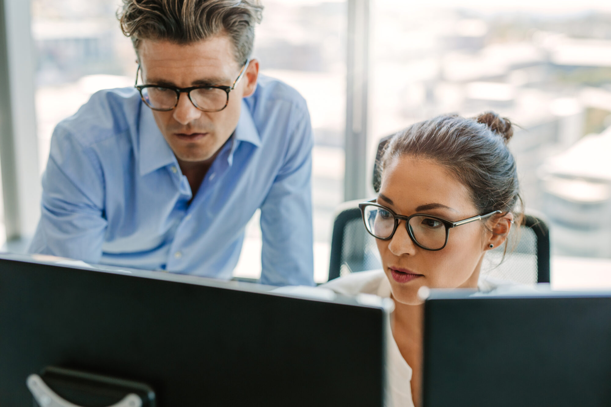 A man and a woman, both wearing glasses, are focused on computer screens in an office setting. The man is standing while the woman is seated, both deeply engaged with Ava Security software displayed on their monitors.