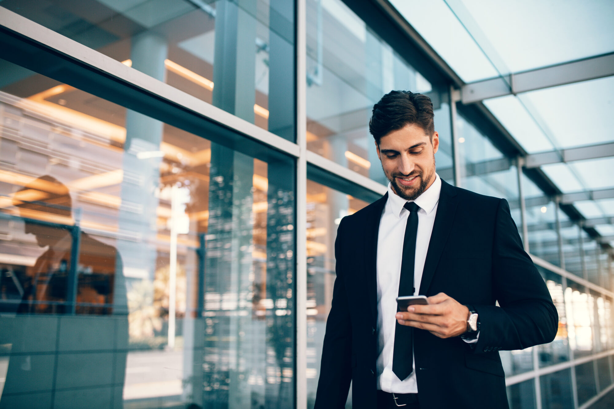 A man in a black suit and tie is walking outside a modern glass building, securely scrolling through his smartphone. He appears to be smiling, the reflections of the cityscape and security integrators' sleek designs visible in the glass panels behind him.