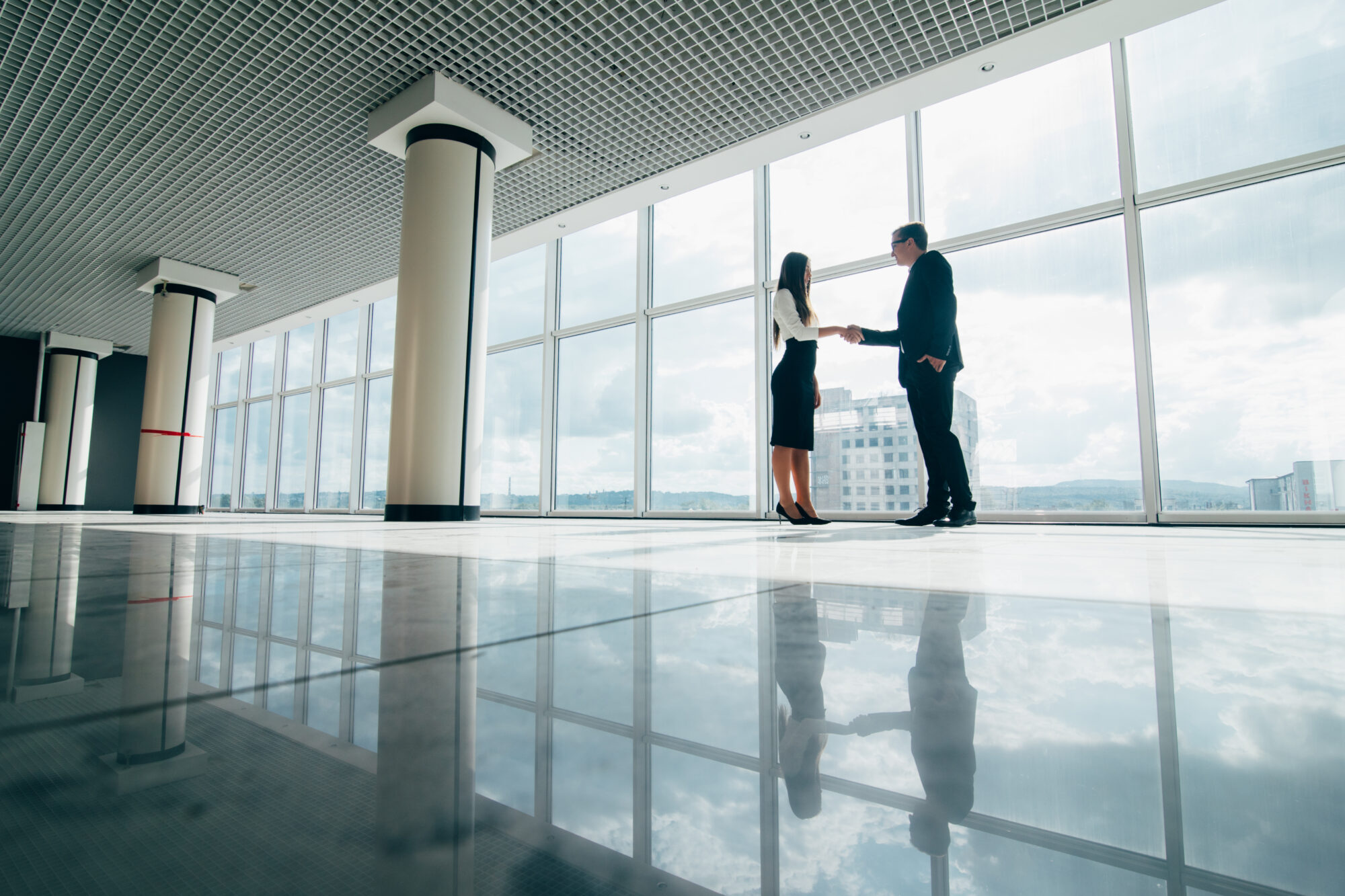 Two people, a man and a woman, are shaking hands in a spacious, modern office with large windows offering a city view. The floor reflects their silhouettes. The room is mostly empty, creating a sense of openness—ideal for discussing seamless property access management solutions.