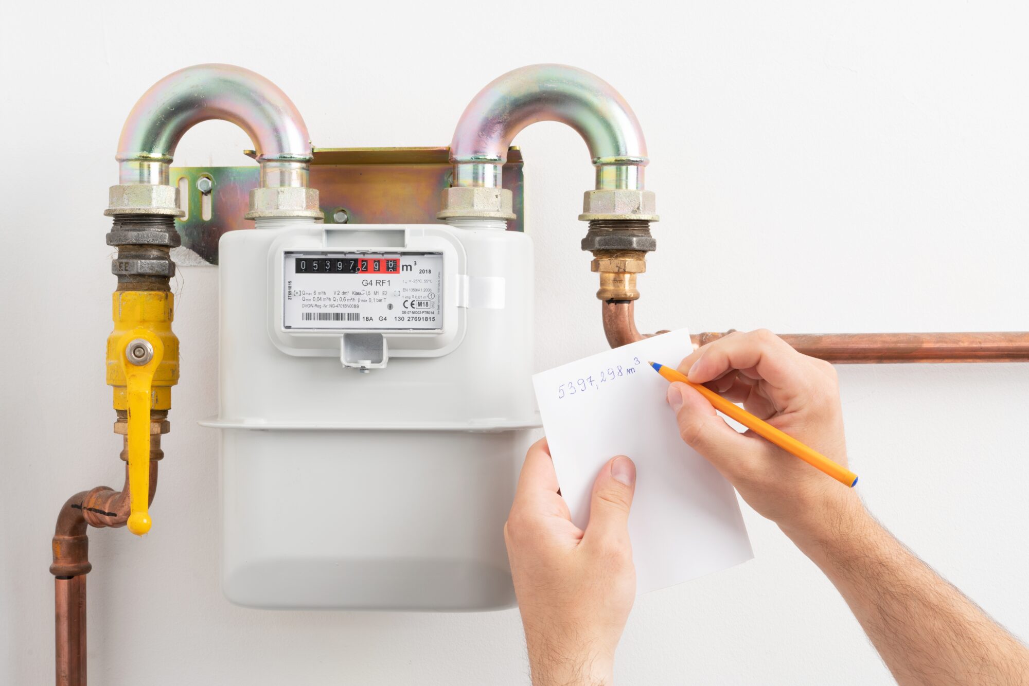 A person records the numbers shown on a residential gas meter with a pencil and paper. The smart gas meter is connected to copper pipes and displays several numbers on its digital screen.