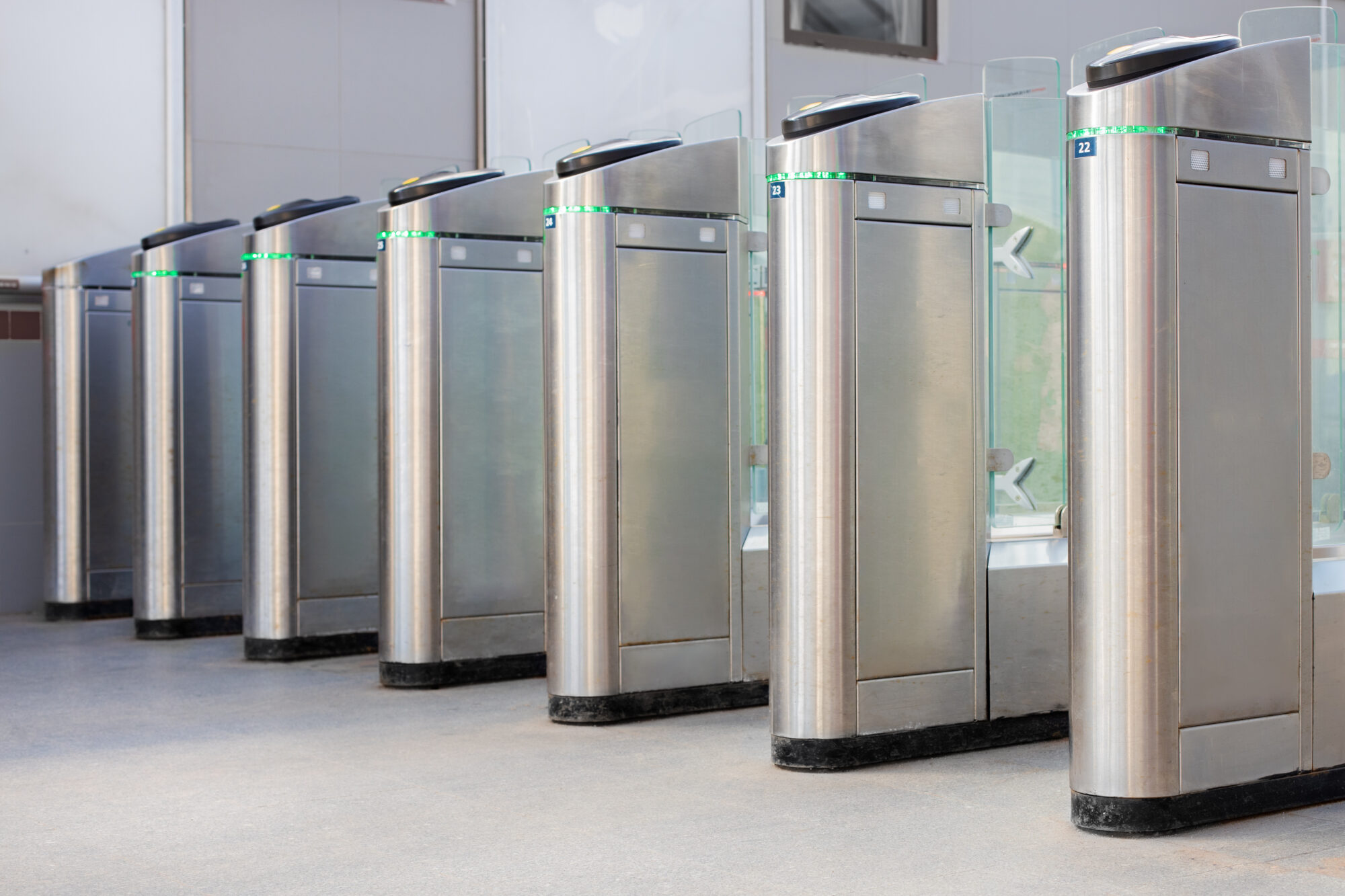 A row of modern, stainless steel automatic turnstiles in a brightly lit entrance area, each featuring a vertical glass barrier and green indicator lights, operates on an attribute-based access control system typically used in transit stations or secure buildings.