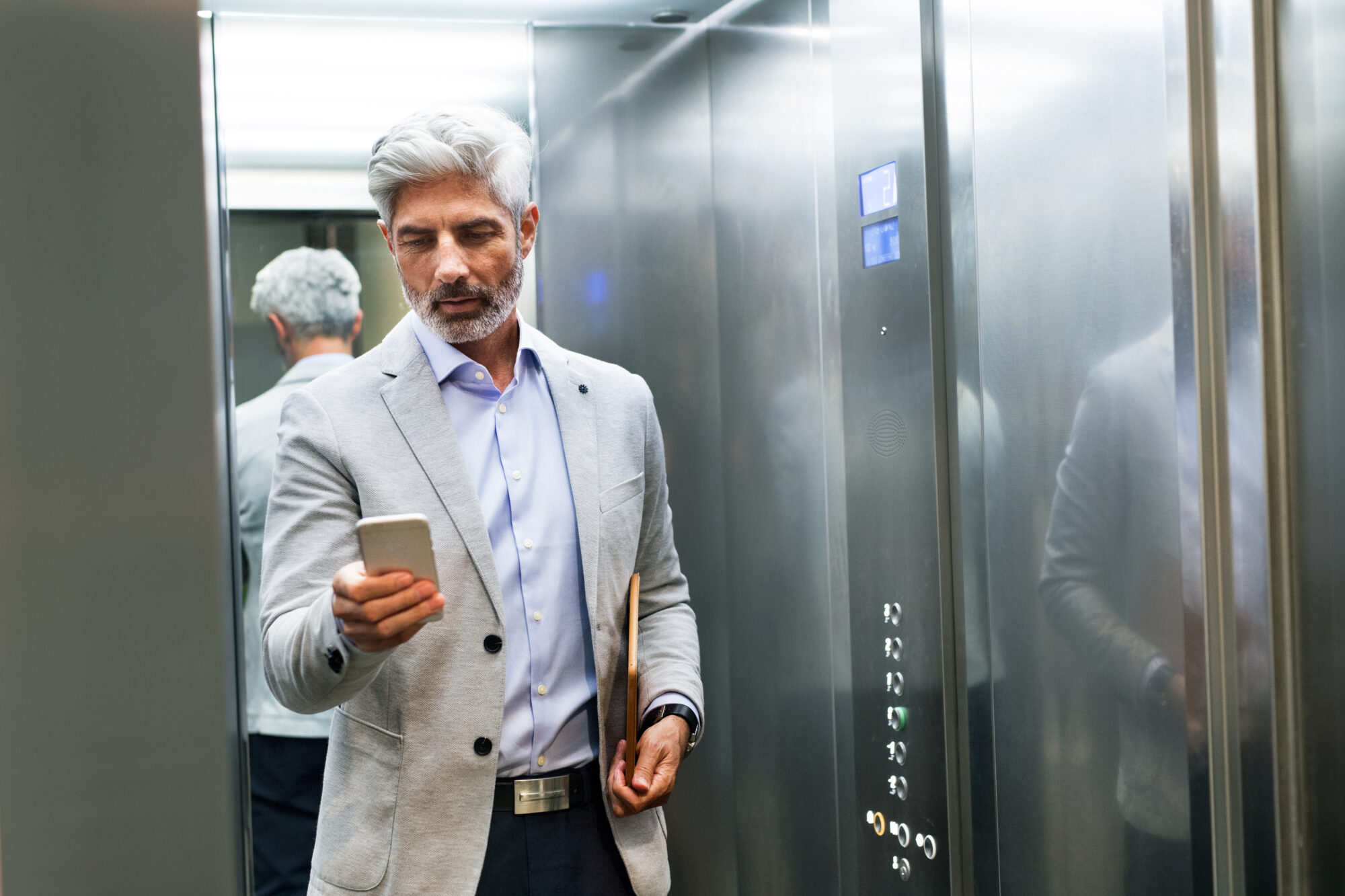 A man with gray hair and a beard, dressed in a light gray suit and light blue shirt, is standing in a touchless elevator. He's looking at his phone while holding a folder in his other hand. The metallic walls around him gleam with a reflective sheen.