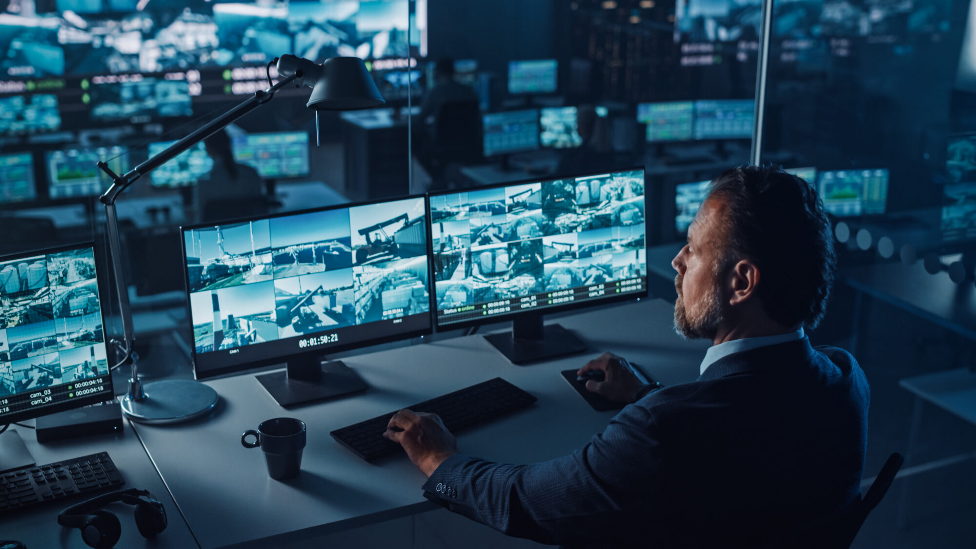 In a dark control room, a person sits focused at a desk, monitoring multiple cloud-based security camera systems on several computer screens. The high-tech atmosphere is accentuated by additional screens and computer equipment visible in the background.