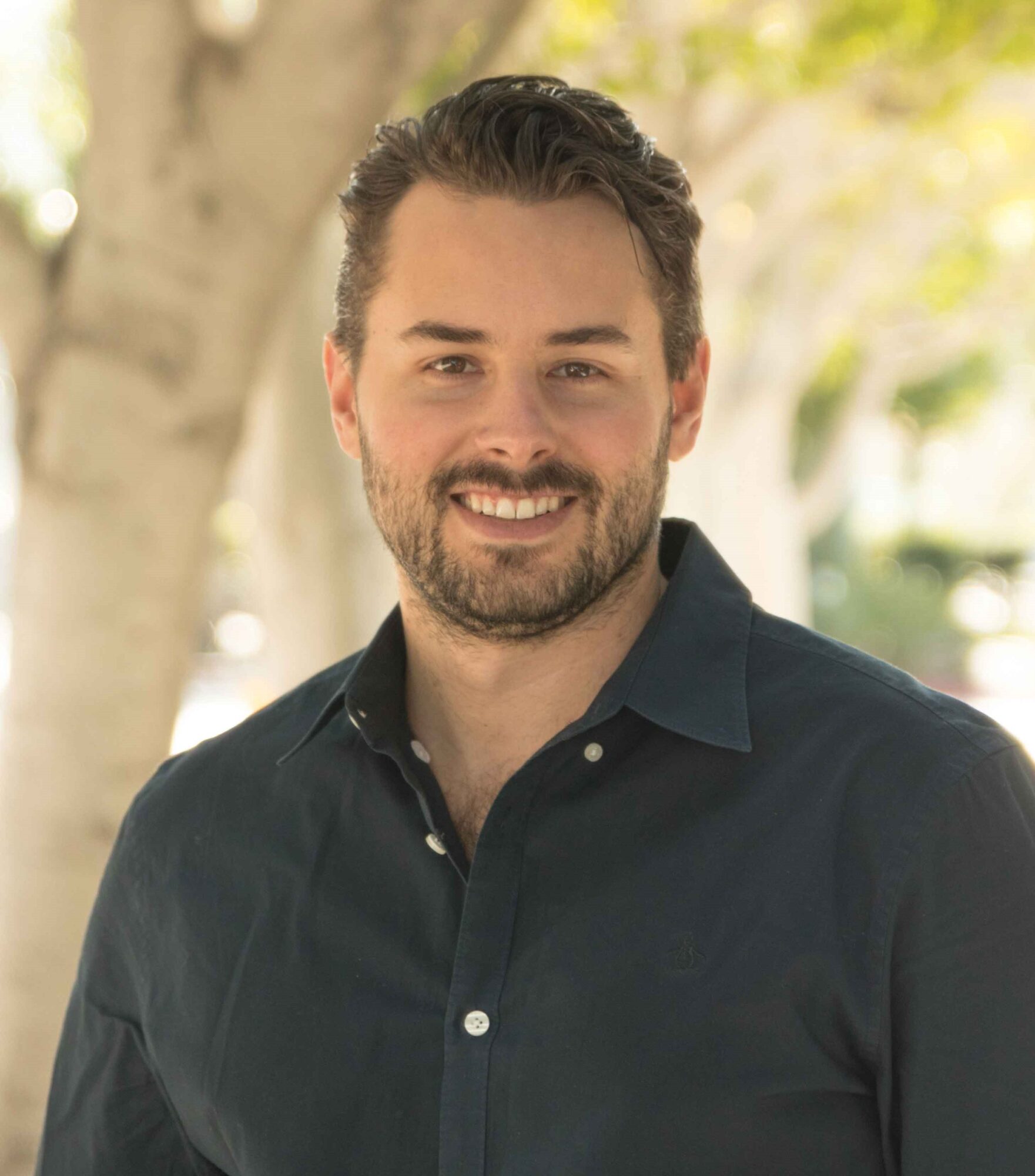 A man with a beard and dark hair smiles while wearing a dark button-up shirt. He stands outdoors, evoking curious thoughts about Genea, surrounded by blurred green trees and vibrant sunlight in the background.