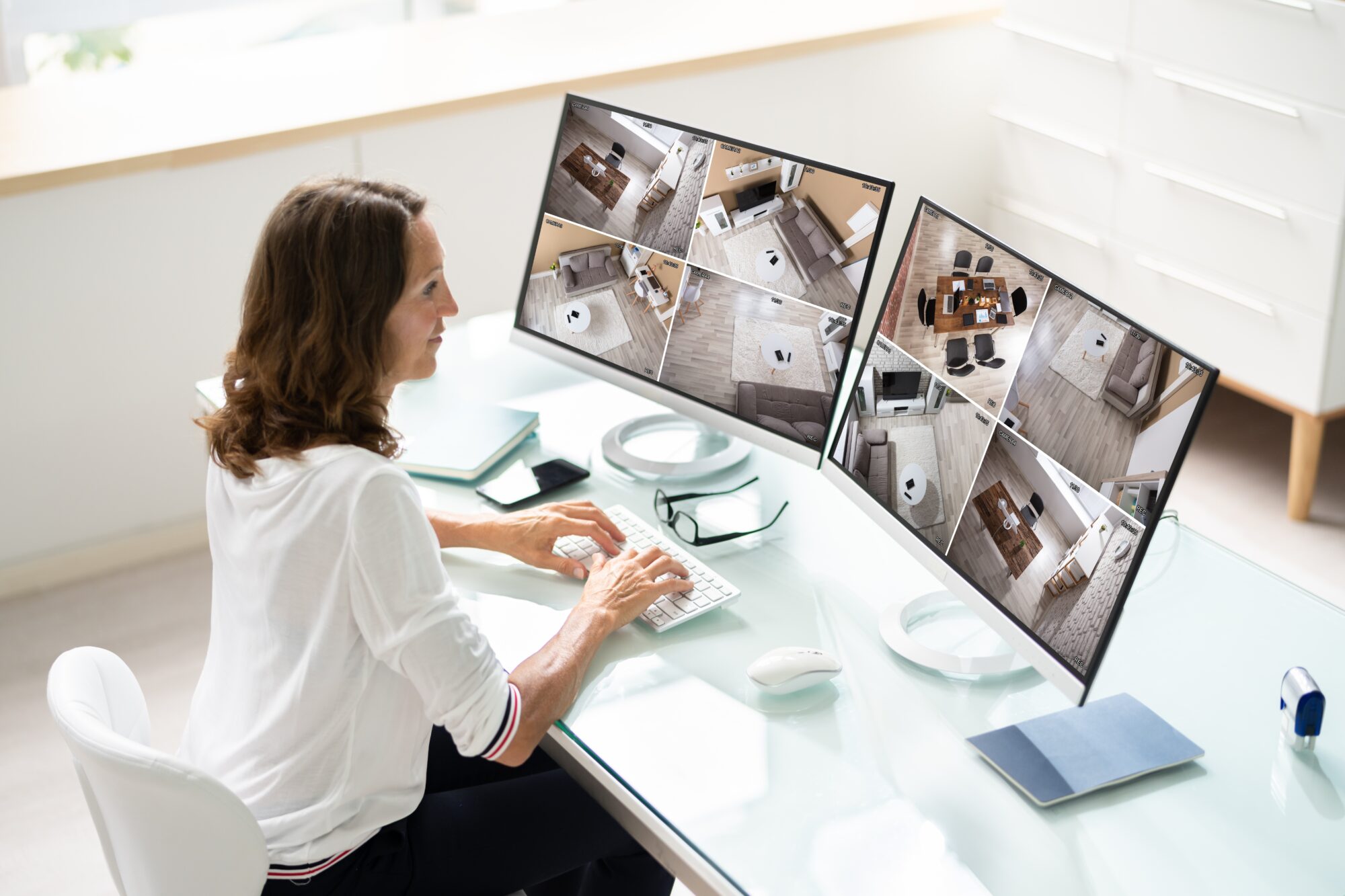 A person is seated at a desk using a keyboard and mouse, attentively managing salient video feeds on two large monitors, showcasing security cameras monitoring various interior rooms. The bright office space is minimalistic and organized.