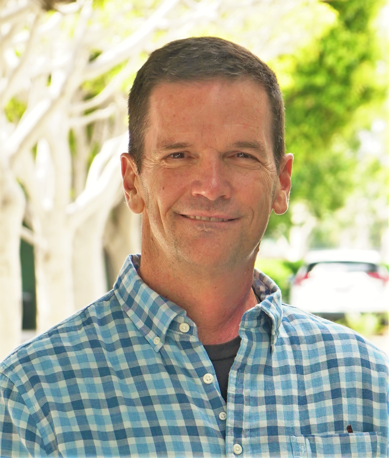 A man in a blue plaid shirt smiles at the camera while standing outdoors, perhaps reflecting on his thoughts about Genea. The background is softly blurred, with trees and a car visible in the distance.