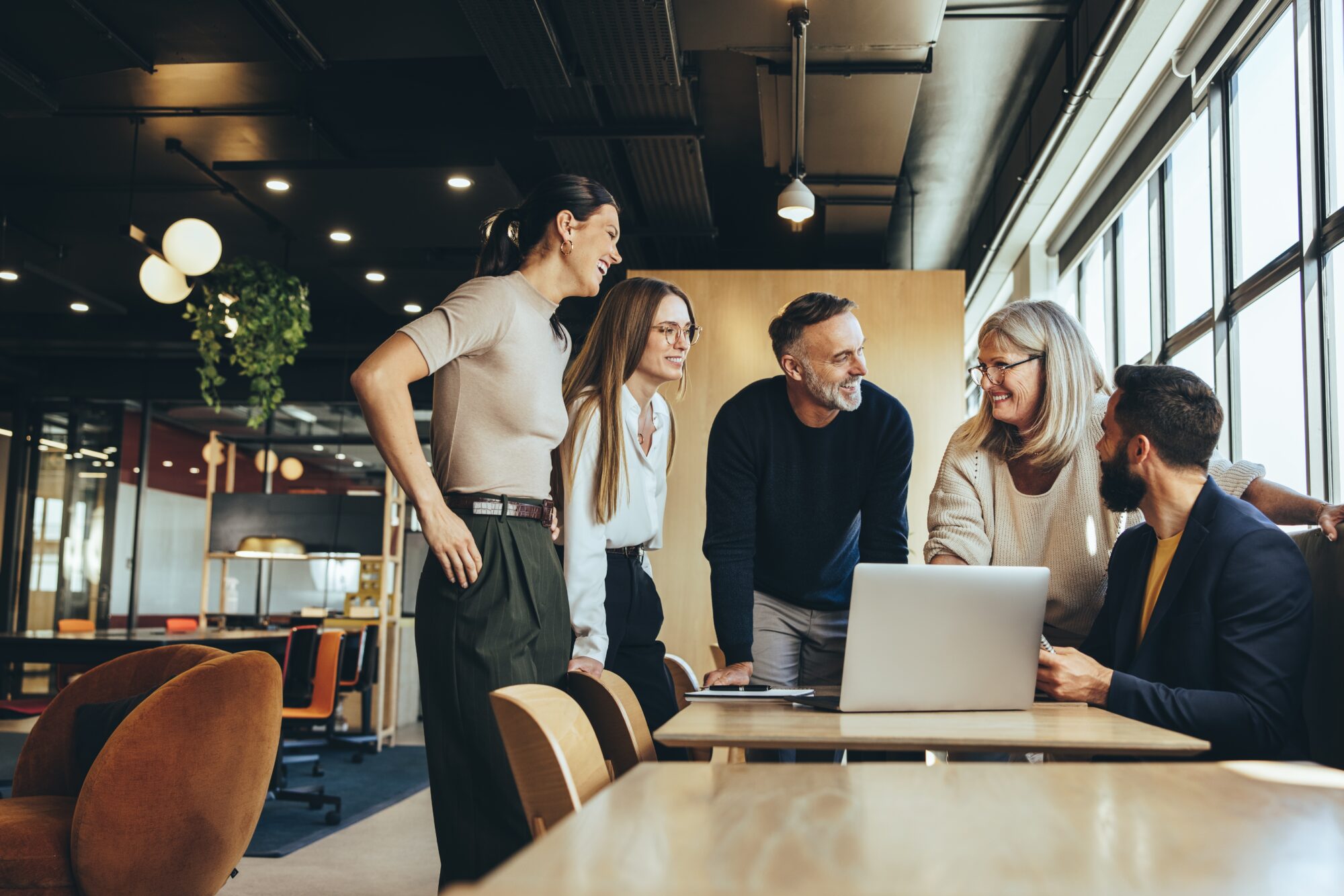 A diverse group of five people in a modern office setting gather around a laptop, engaged in a positive Webex discussion. Large windows to the right let natural light illuminate the space, which features sleek furniture and greenery.