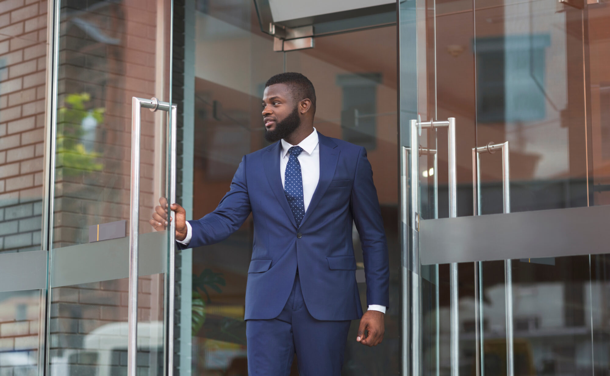 A man in a blue suit, white shirt, and dotted tie is opening an Allegion glass door to a building. He has a beard and looks to the side with a focused expression. The building features brick and glass elements.