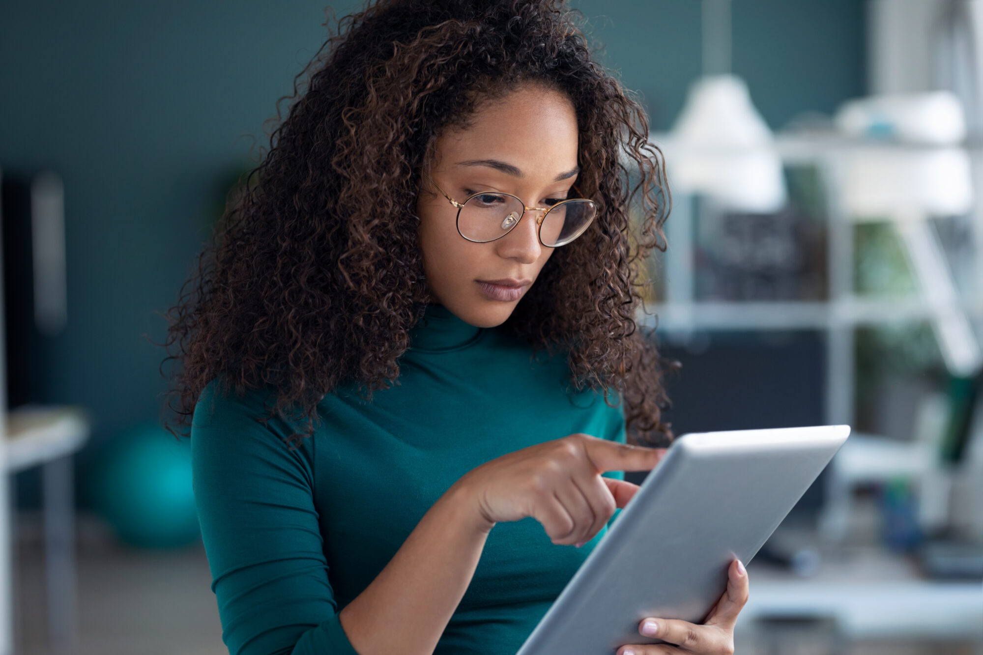 A person with curly hair and glasses is standing indoors, intently reviewing access logs on a tablet with one hand. They are wearing a green top, and the background includes blurred indoor elements like lights and furniture.