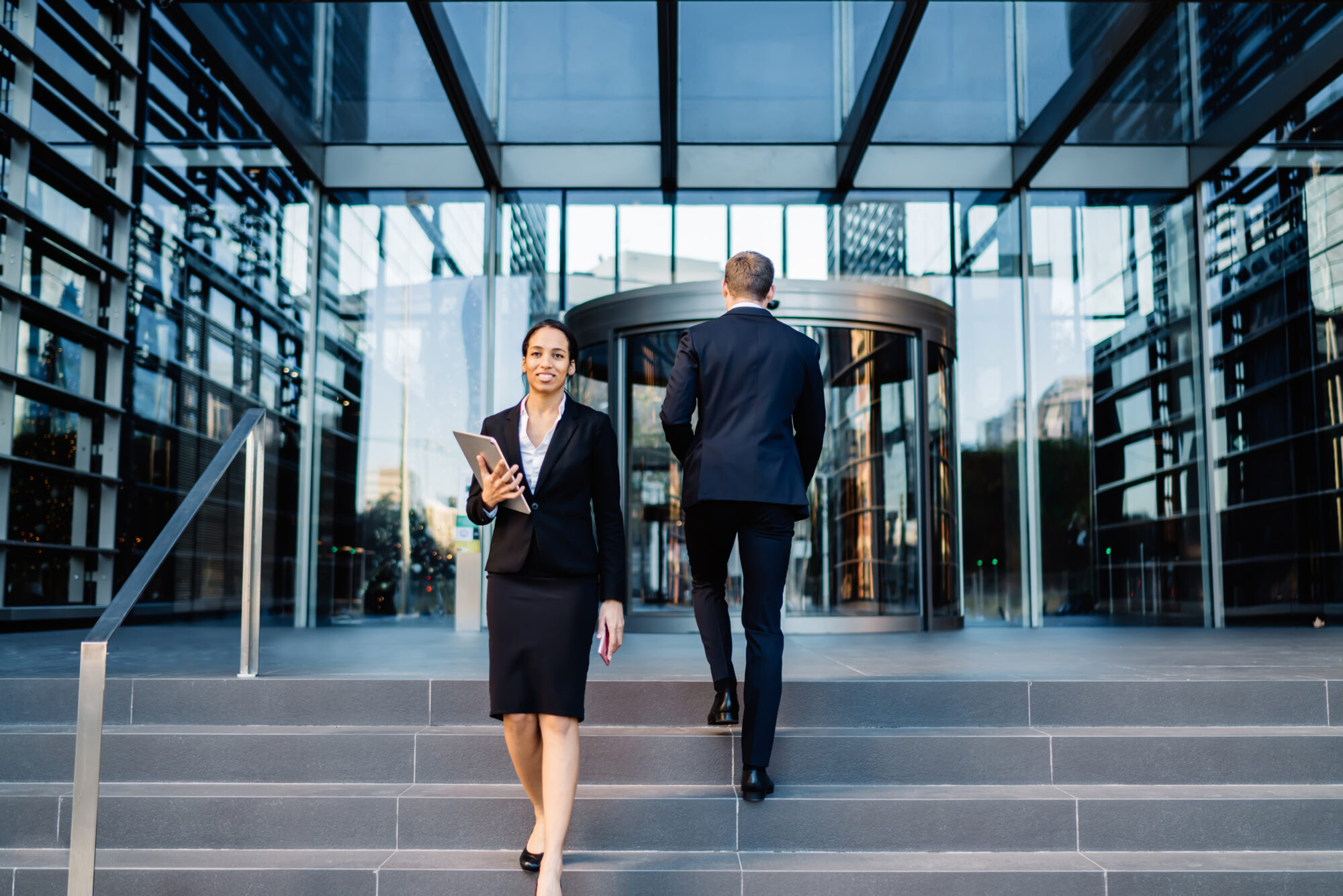 A woman in business attire, holding a tablet, walks down steps in front of a modern glass building. A man in a suit approaches the entrance, where seamless access control ensures security. The scene is set in an urban environment.