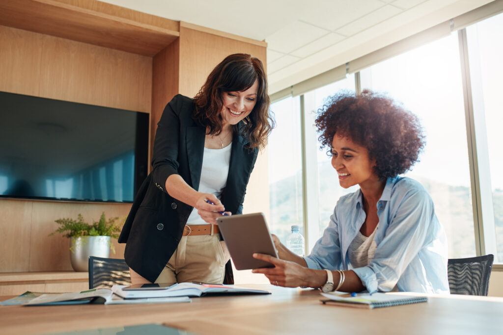 Two women in a bright office setting engage in a discussion. One stands, smiling, gesturing at a tablet displaying interactive floor plans held by the seated woman. A large screen and window form the backdrop of their collaborative moment.