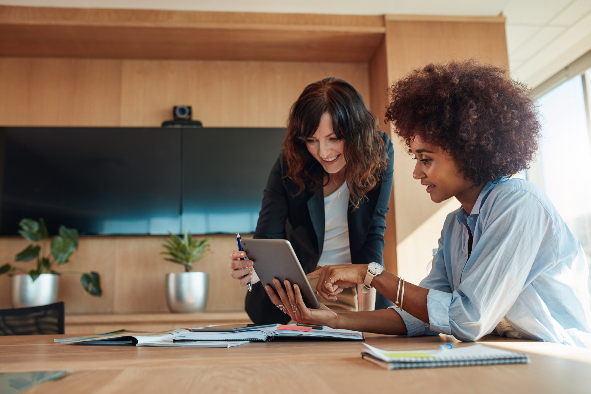 Two women in a modern office setting examine a tablet together, exploring interactive floor plans. One stands while the other is seated, surrounded by notebooks and documents on a wooden table. A large monitor and potted plants create a dynamic backdrop.