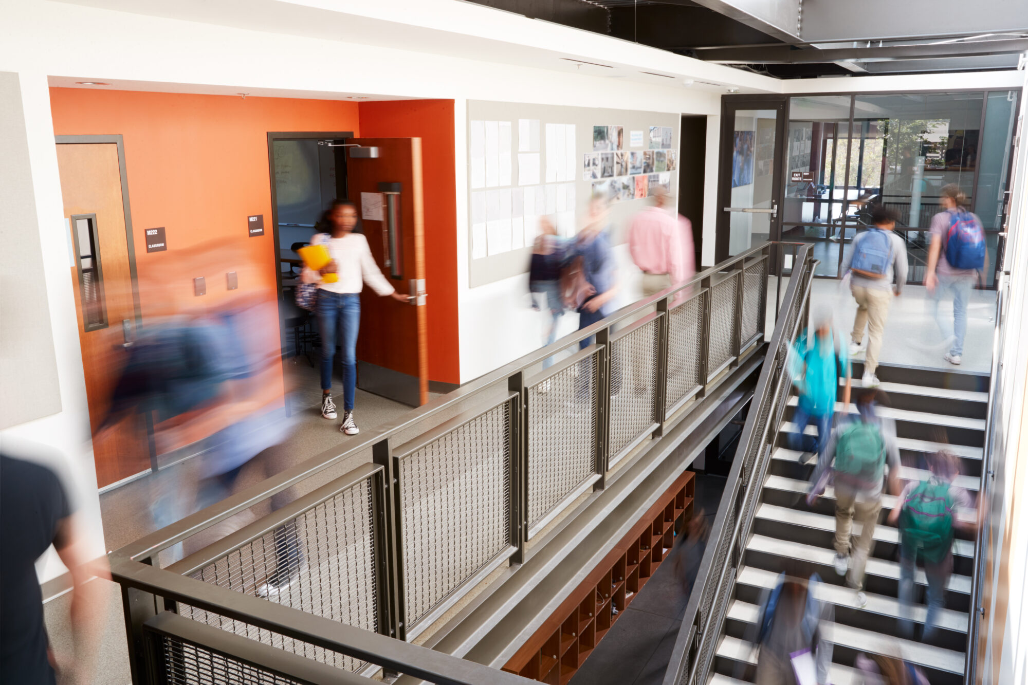 A busy school corridor with students walking past classrooms buzzes with energy. Some head up the staircase to another level, captured in motion blur. The scene underscores a modern school environment committed to safety measures like Alyssa's Law, enhancing security and peace of mind.