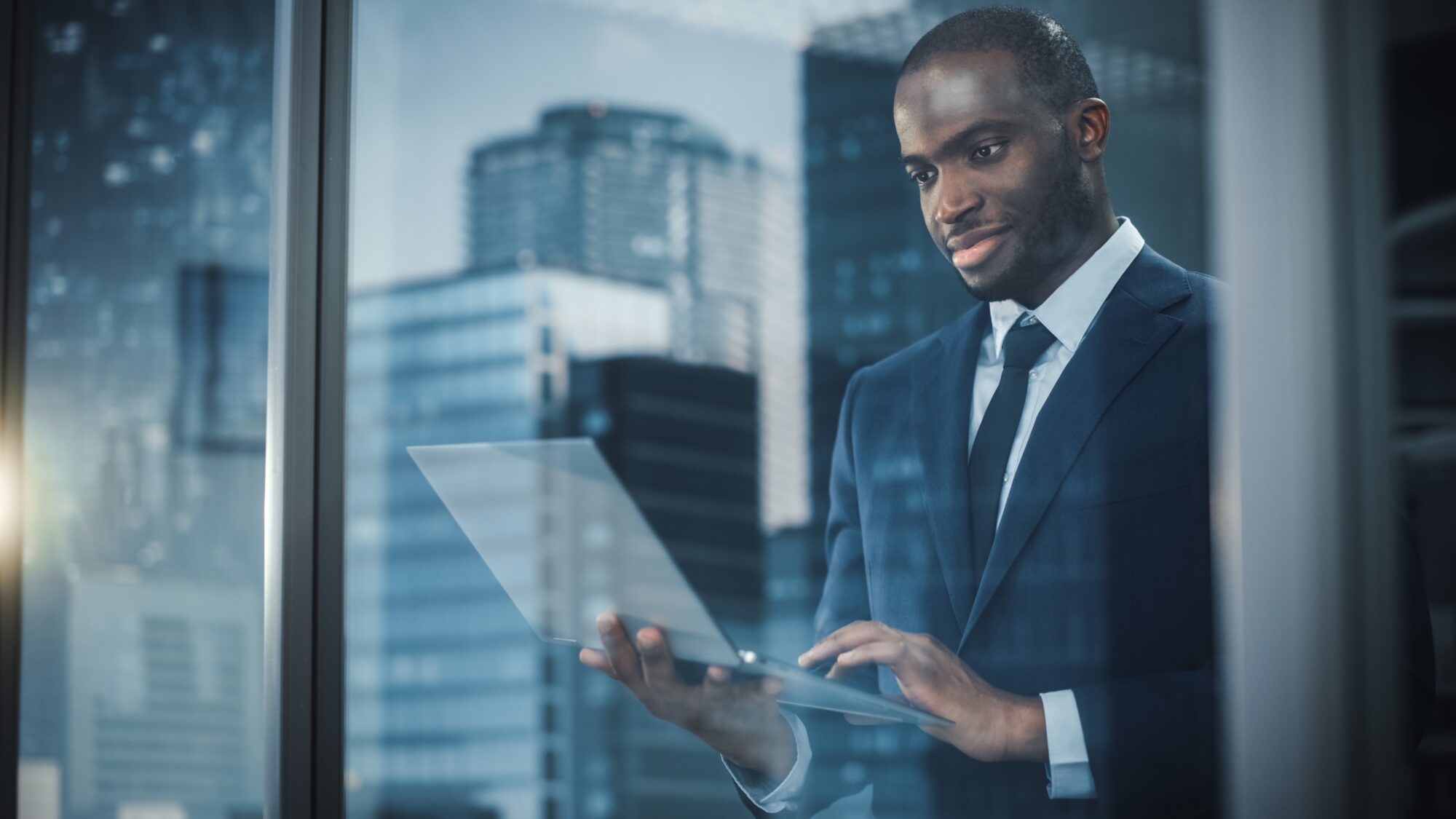 A man in a suit stands by a window, holding a laptop in a modern office. The cityscape with tall buildings is reflected clearly on the single pane of glass, enhancing his focus and engagement with his work.