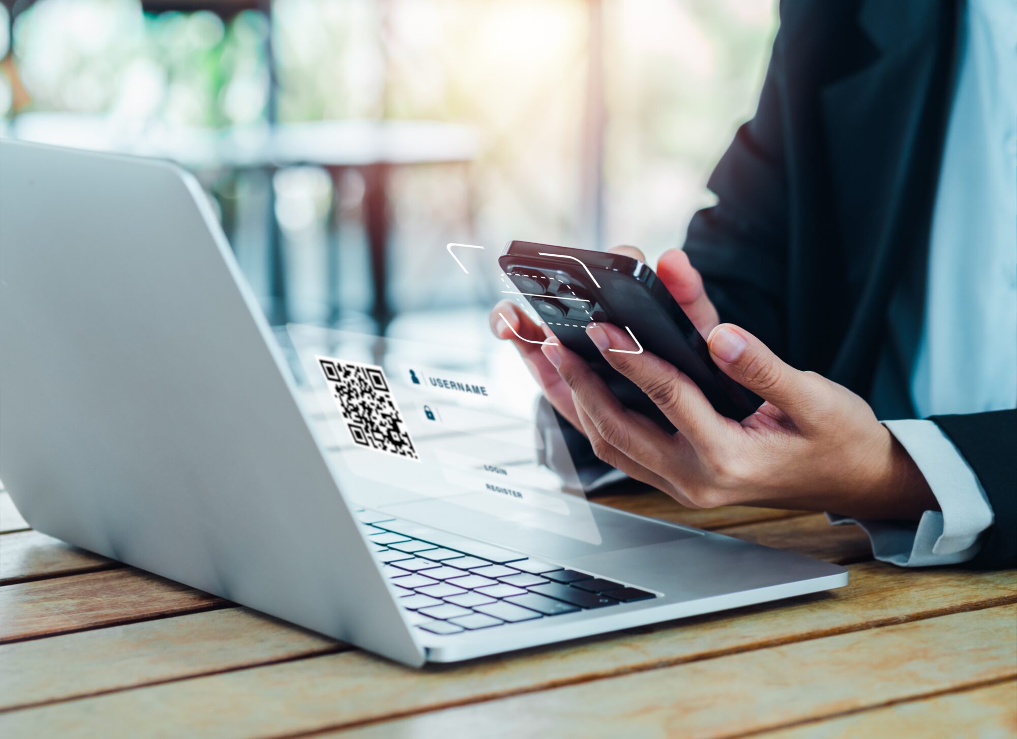 A person scans a QR code for access control from a laptop screen using their smartphone. The background is blurred, and the setting appears to be a casual workspace with natural lighting.