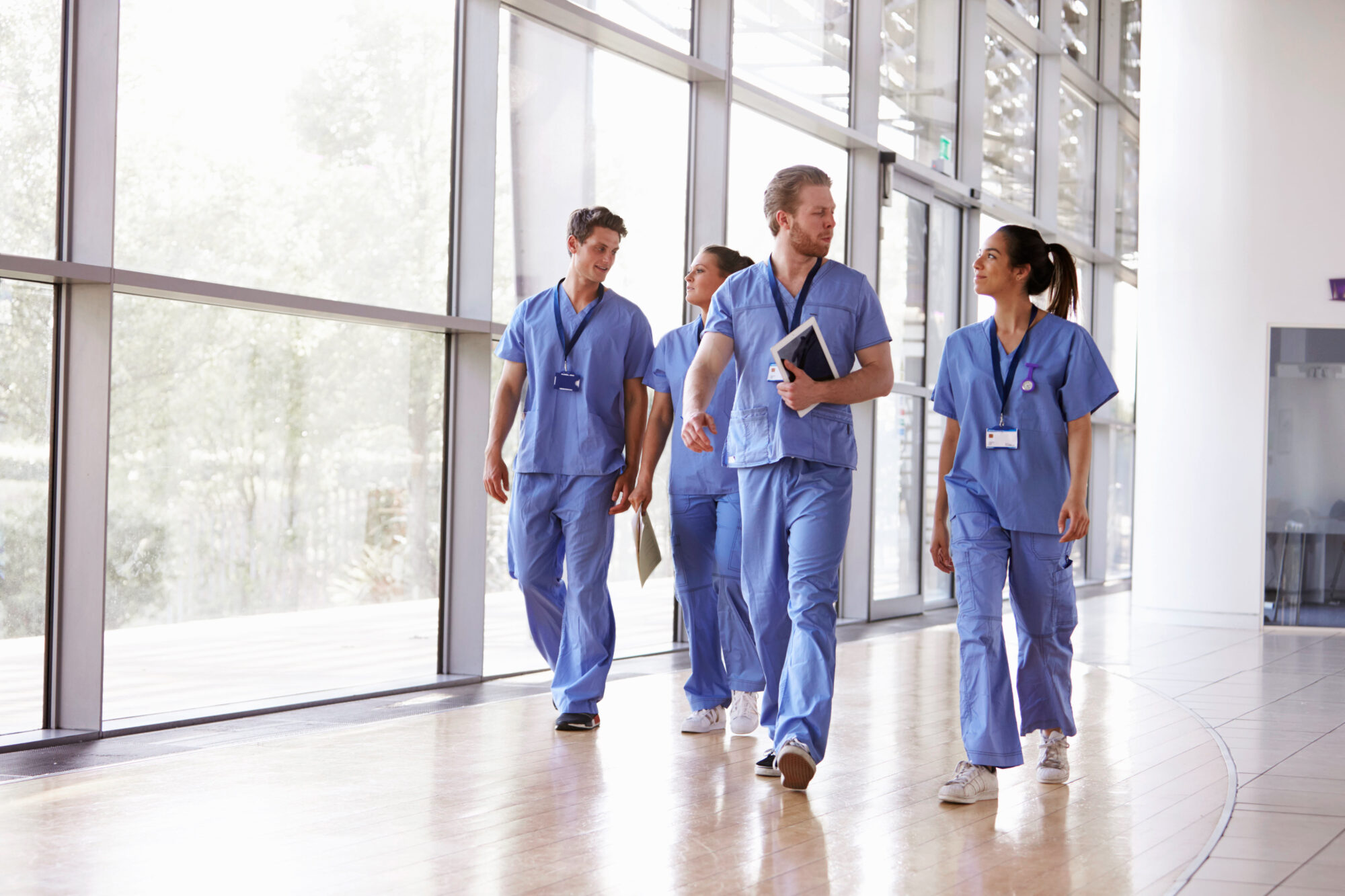 A group of healthcare professionals in blue scrubs walks down a sunlit hallway, engaged in conversation about access control for hospitals. They hold notepads and folders as large windows line the corridor, allowing natural light to illuminate the space.