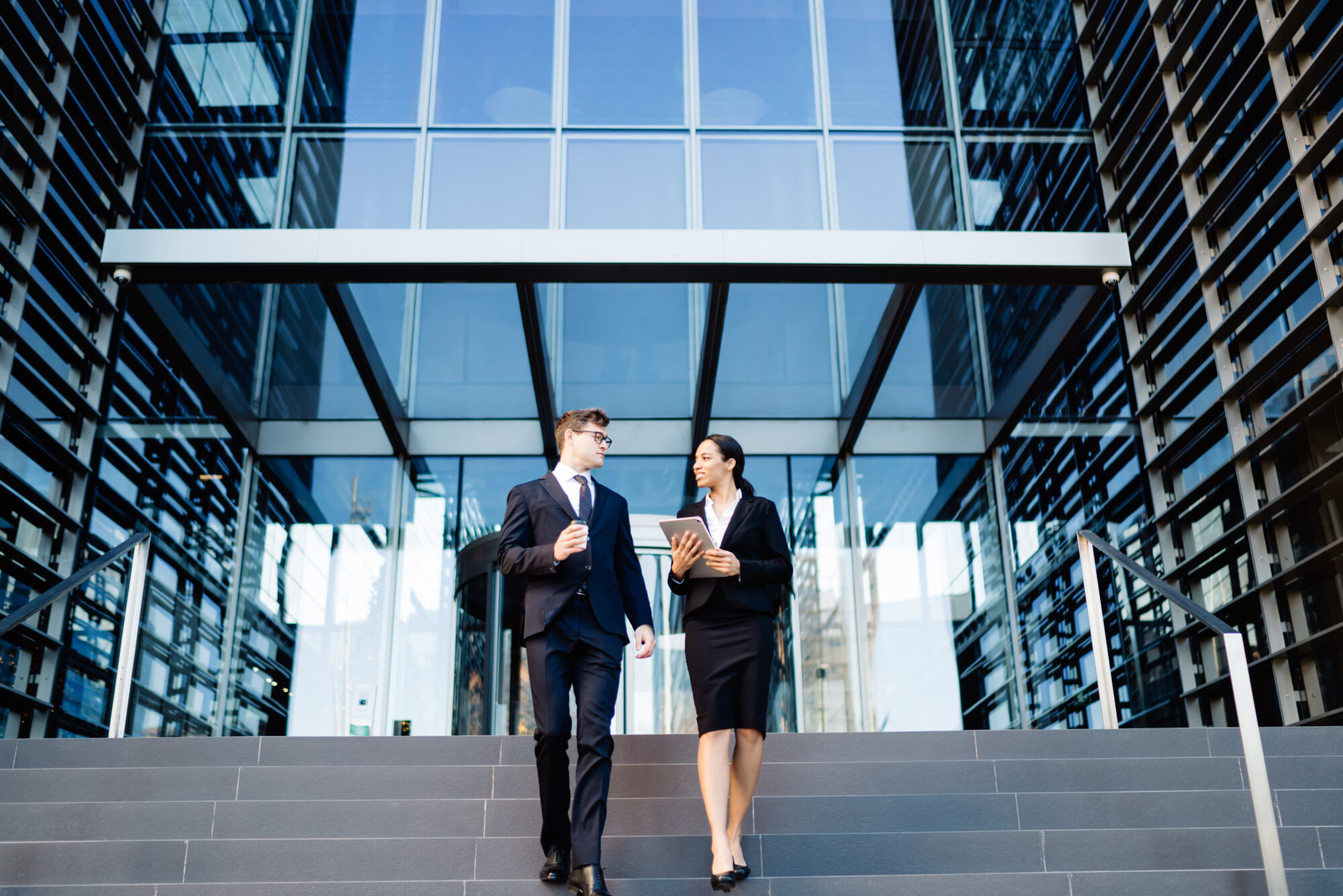 Two professionals in business attire walk down steps from a modern glass building, engaged in a conversation about enterprise access control. The man carries a coffee cup while the woman holds a tablet. The setting suggests a corporate or office environment.