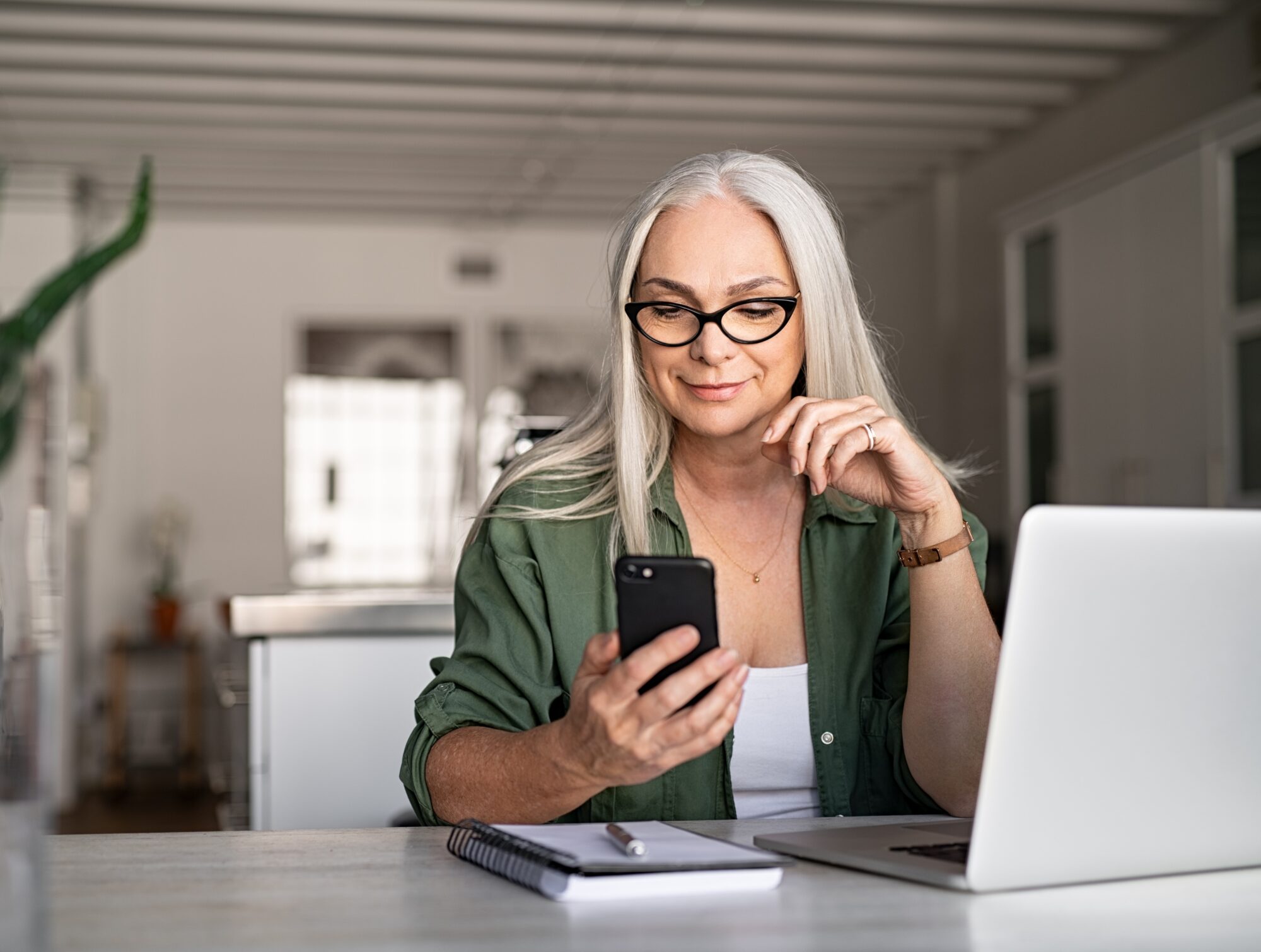 A woman with long gray hair and glasses is sitting at a table, looking at her smartphone. In the bright room, surrounded by her laptop and notebook, she appears focused and relaxed as she reviews submeter billing details.