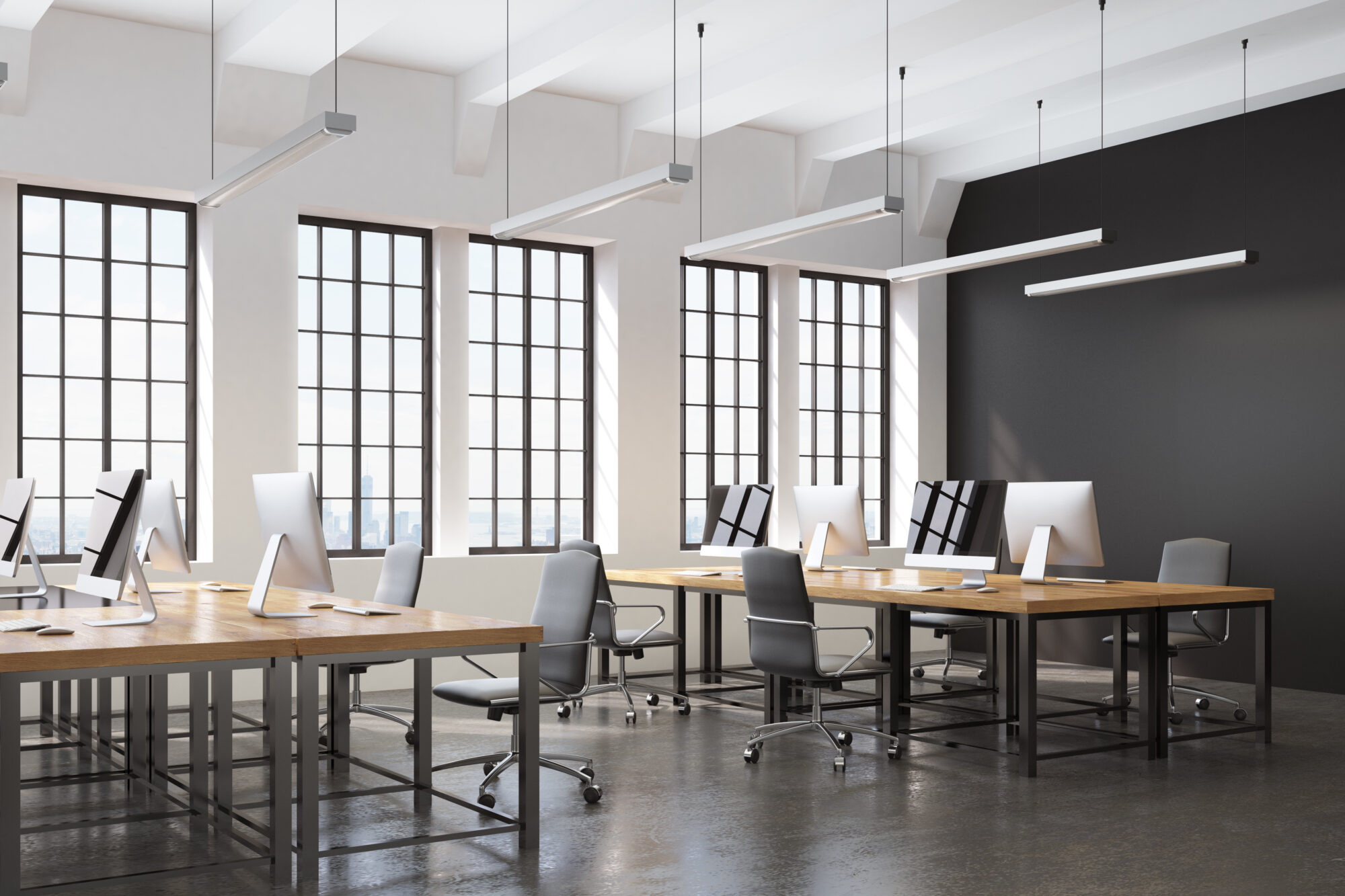 Modern office space with large windows, wooden desks, and multiple computer workstations. Gray chairs are neatly arranged at each desk. The room, perfect for reviewing holiday schedules for access control, boasts a clean, minimalist design with overhead lighting and a dark accent wall.