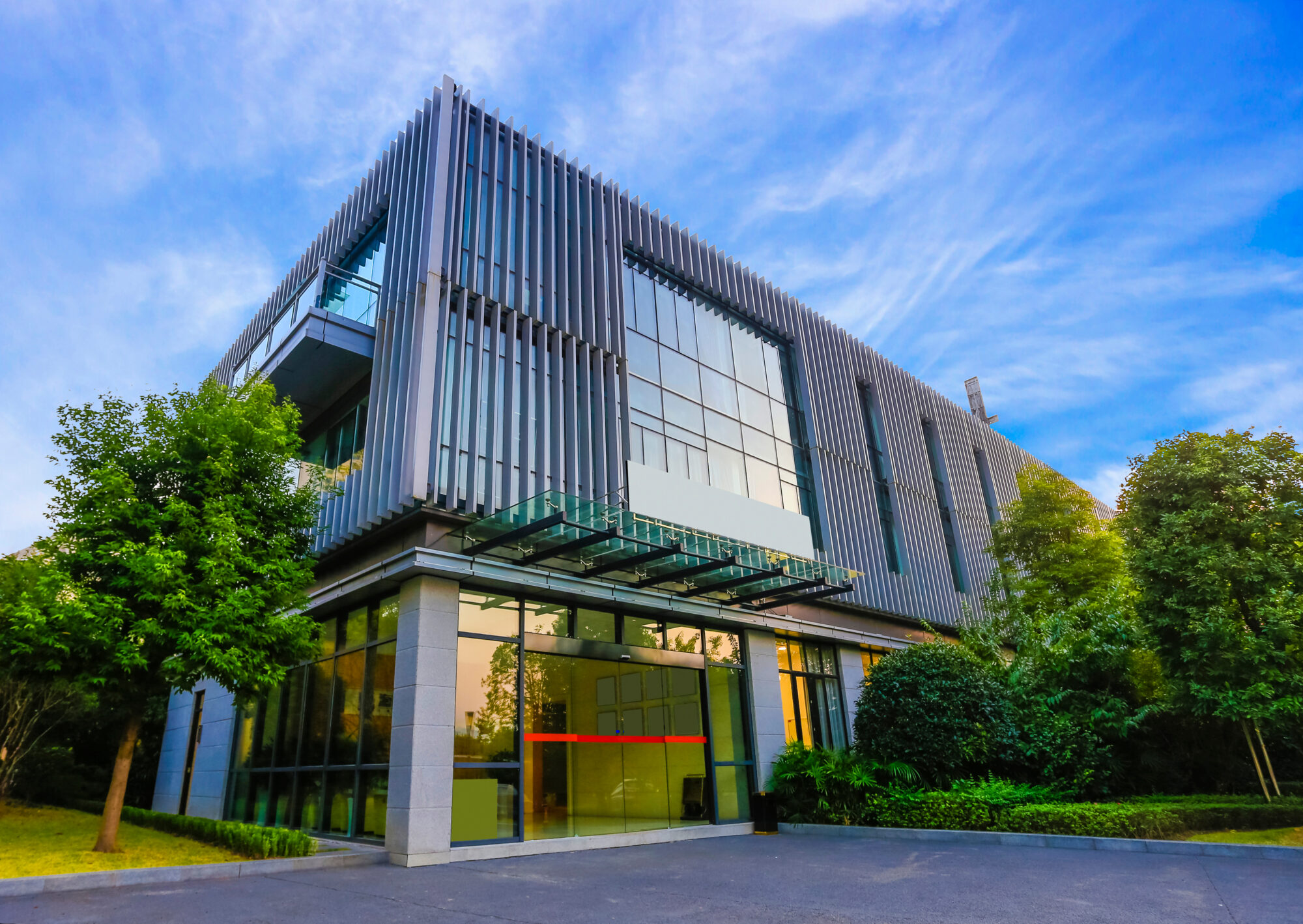 Modern two-story building with large reflective windows and vertical metal paneling, set against a clear blue sky. Equipped with commercial submetering, it is surrounded by green trees and bushes, with a paved driveway leading to the entrance.