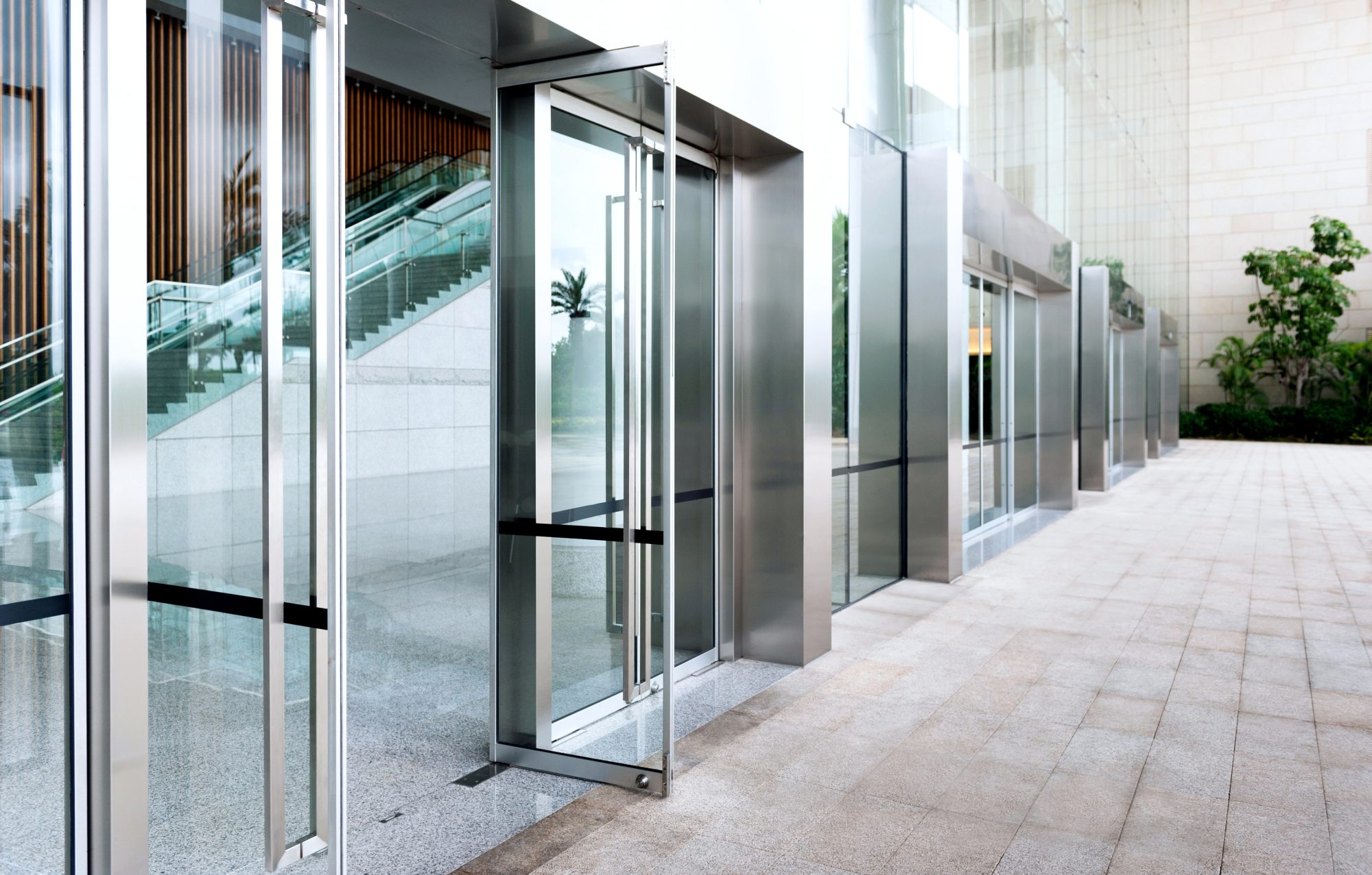 A row of modern glass doors with metal frames, equipped with access control, opens to a large, bright entrance. An indoor escalator is visible in the background, and there are plants and a light stone wall along the exterior walkway.