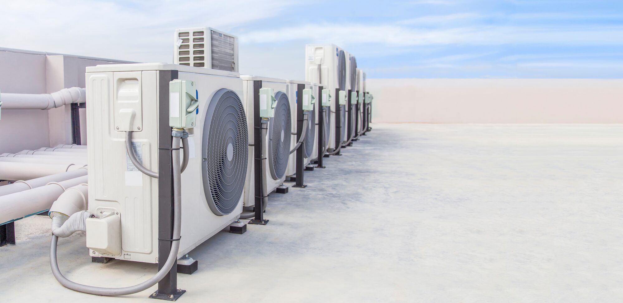 A row of outdoor air conditioning units is positioned on a flat rooftop under a clear blue sky. The units, part of an efficient HVAC system, have large fans visible on their sides and are connected by various pipes and cables, helping to manage HVAC costs effectively.