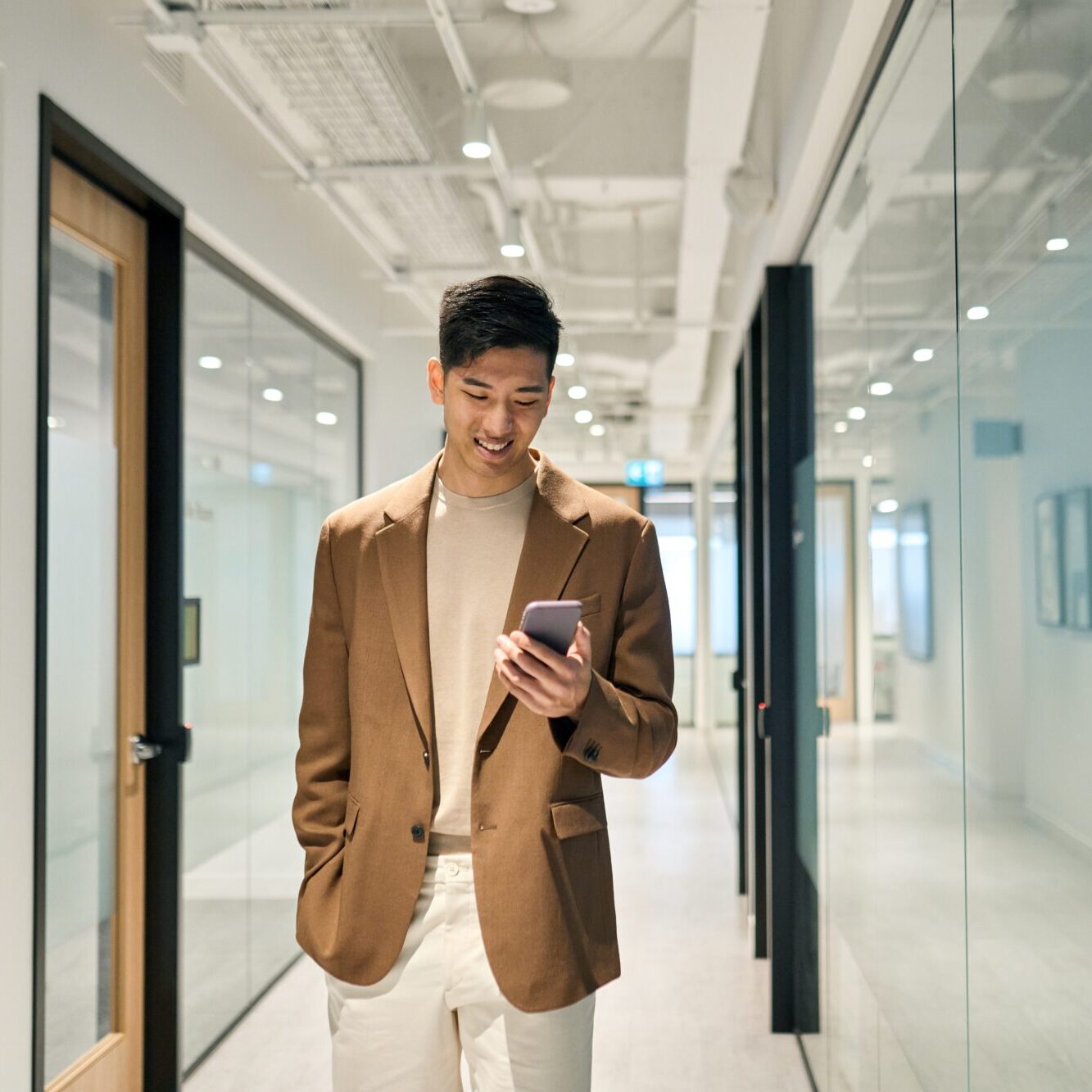 A person in a brown blazer and white pants stands in a modern office hallway, checking Google Wallet on their smartphone and smiling. The corridor features glass walls and doors, illuminated by bright overhead lighting.