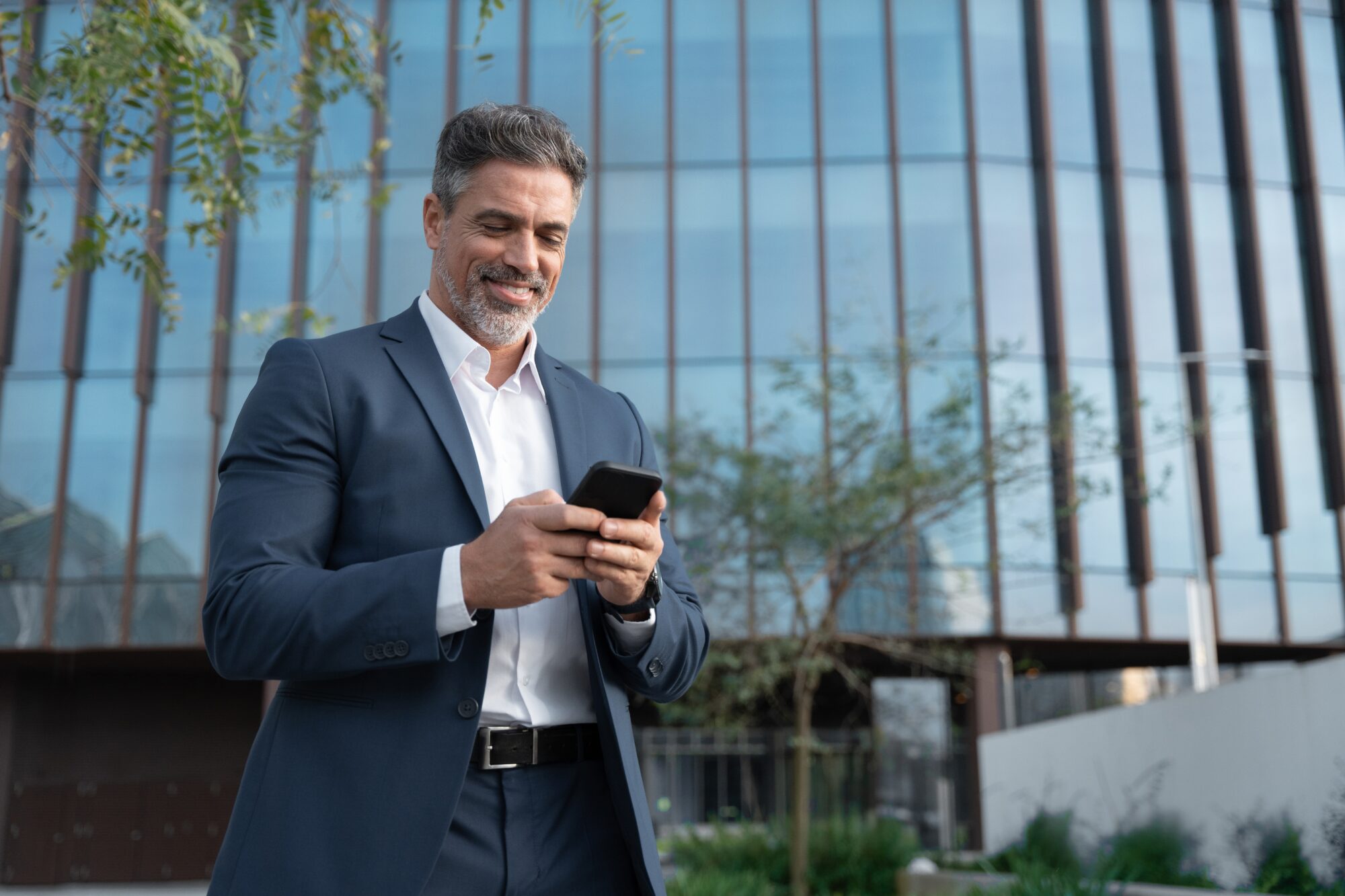 A man in a dark suit smiles while checking Google Wallet on his smartphone. He stands outdoors in front of a modern glass building, surrounded by trees and greenery. The sky is clear, suggesting a bright day.