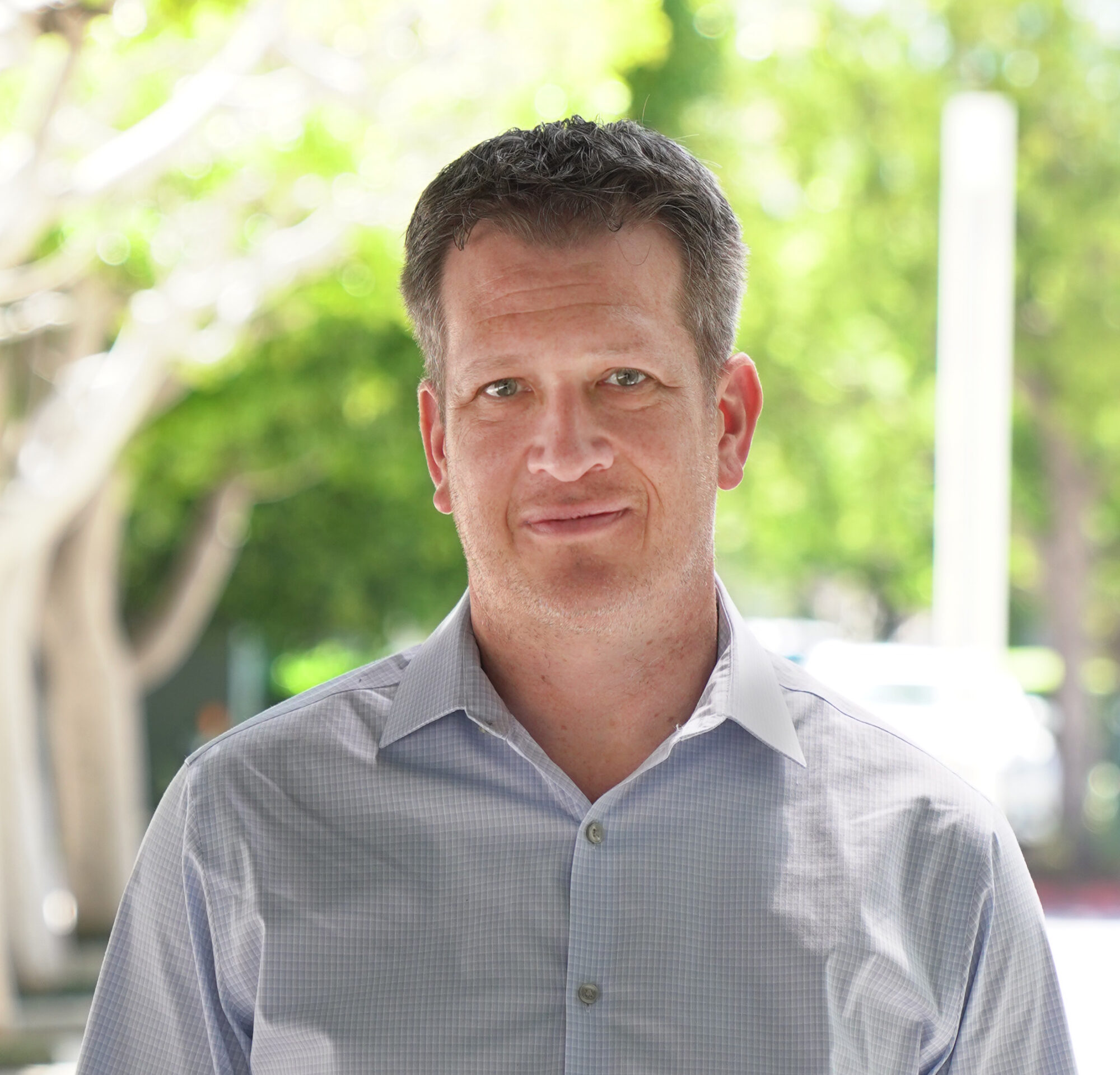 A man with short hair, wearing a light gray button-up shirt, stands outdoors on a sunny day. The background is blurred, with green trees and a pathway leading to discussions about Genea's impact on the environment.