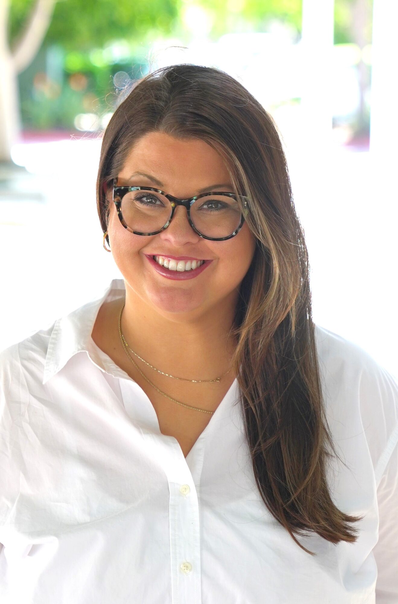 A smiling woman with long brown hair and glasses, embodying the essence of Genea, is wearing a white shirt. She stands outdoors, framed by a backdrop of lush green foliage.