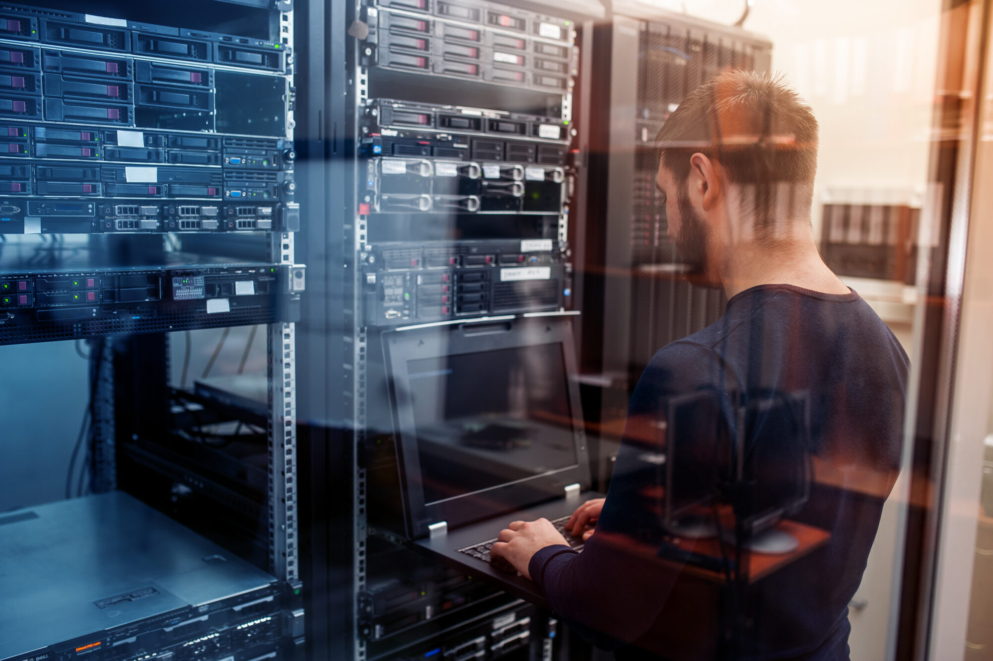 A man stands in a server room, working on a laptop perched on a server rack. Surrounded by rows of servers, their lights flicker like tiny realms in orbit. The scene is reflected in a glass panel, capturing the mercury-like fluidity and complexity of data flow.