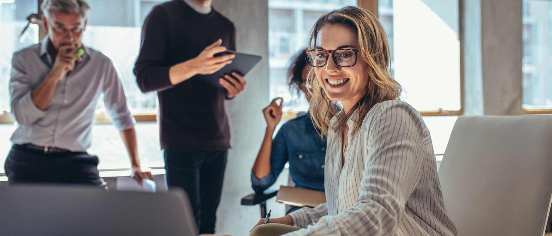 A group of four colleagues is gathered in a bright office. A smiling woman wearing glasses sits in the foreground, as the others stand or sit nearby, engaged in discussion, utilizing a tablet and writing materials to explore new tenant billing software.