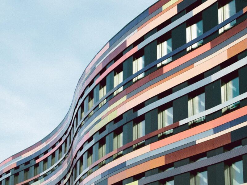 A modern building with a wavy facade features colorful horizontal stripes in red, pink, orange, and blue. Its grid-patterned windows shimmer against the sky, seamlessly blending innovative design with networked technology for precise manual meter reading.