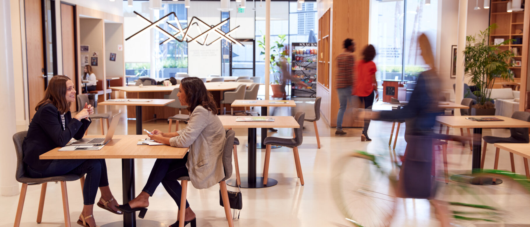 A modern office buzzes with activity as two women chat at a table. Others walk by, while one rides a bike indoors. The bright and open space, enhanced by large windows, plants, and a magazine rack, features advanced mobile access control for seamless entry and security.