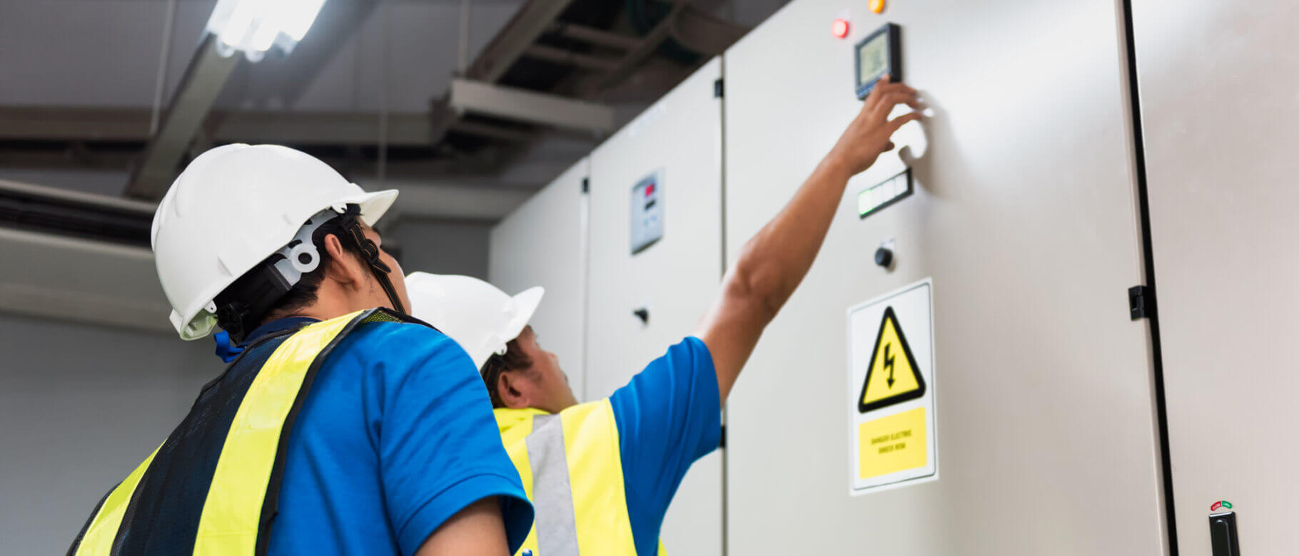 Two workers wearing white hard hats and reflective vests operate a networked control panel in an industrial setting. One points at the display while the other observes. A warning sign with a lightning symbol is visible on the panel, highlighting the need for precision and safety.