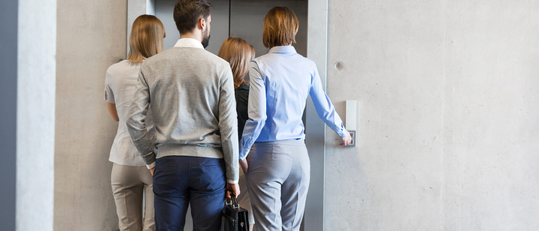 A group of four people, wearing badges and business attire, stands in front of an open elevator door. One person is pressing the button inside. The scene is set against a plain, gray wall, suggesting a modern office environment.