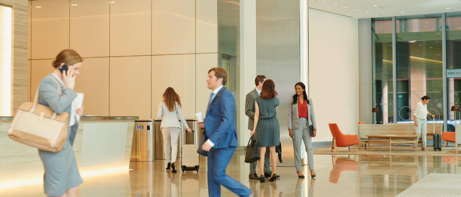 A modern office lobby with people walking and talking. A woman on the left is on her phone, while others walk or converse in groups. Wireless locks secure the entrances, blending seamlessly into the sleek design. Lounge chairs and tables line the right side, against shiny floors and beige walls.