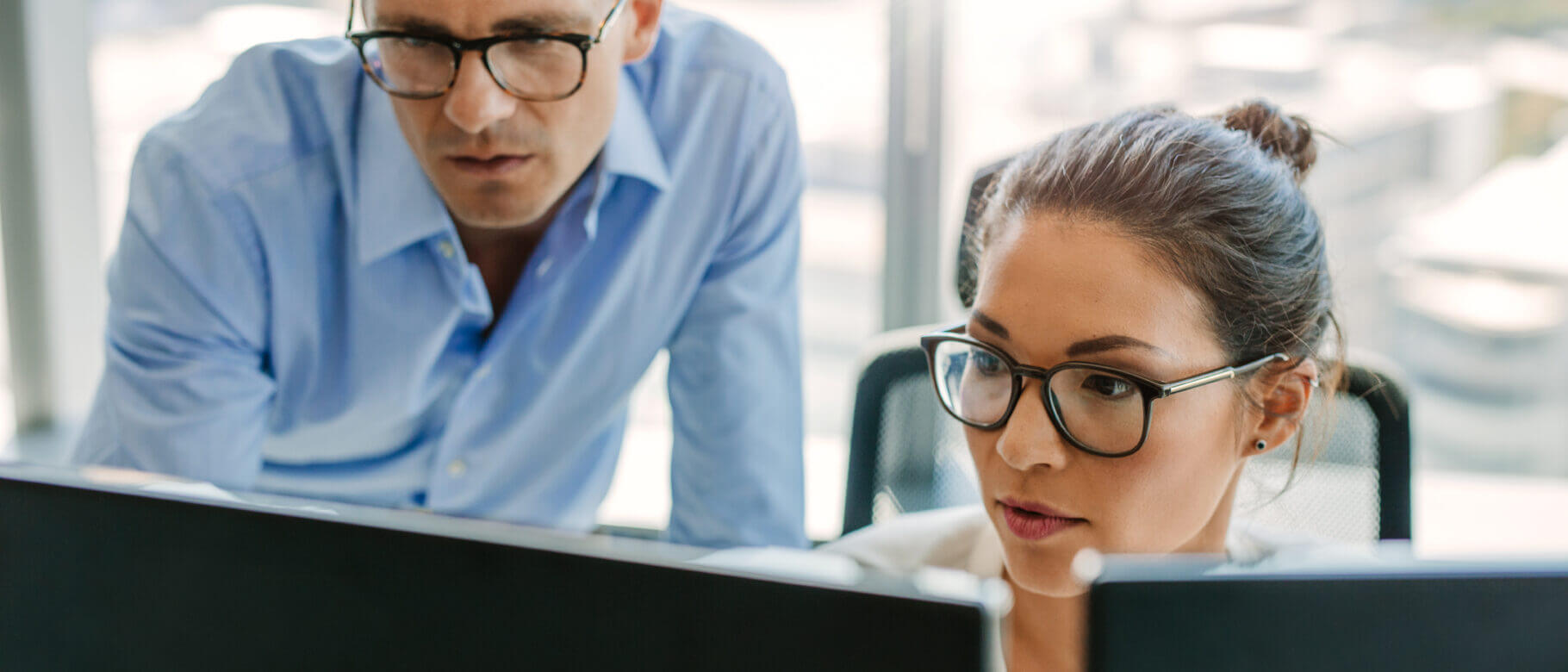 Two people in an office setting focus intently on computer screens. Both wear glasses. The man, dressed in a light blue shirt, leans over while standing, discussing custom roles with the woman who has tied-back hair and sits in front, typing or navigating.